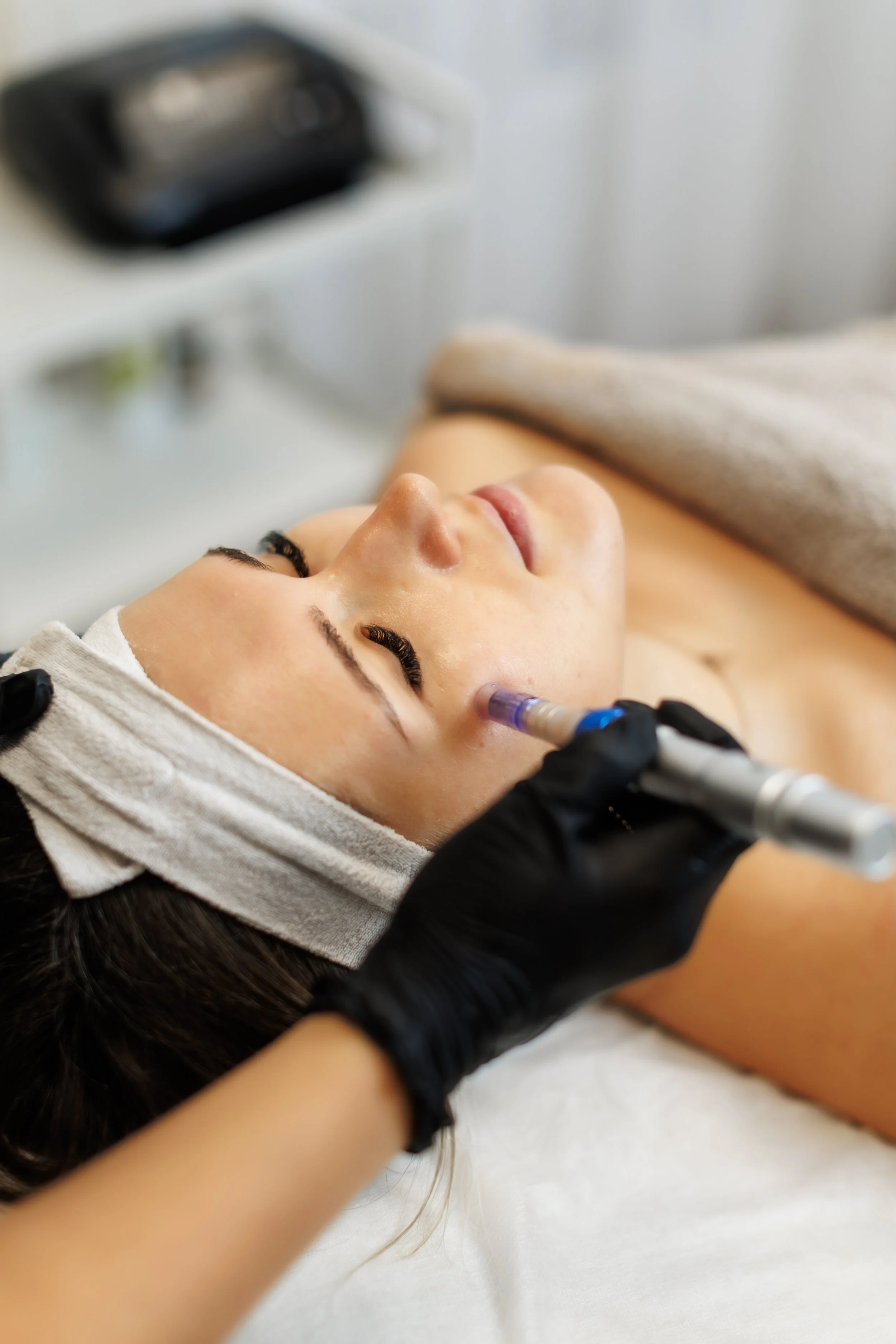 A woman receiving a facial treatment with a syringe-like device on her cheek, lying down with a towel wrapped around her head.