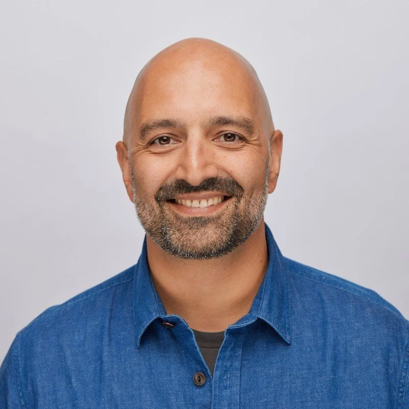 Portrait of a bald man with a beard, wearing a blue button-up shirt, smiling in front of a plain gray background.