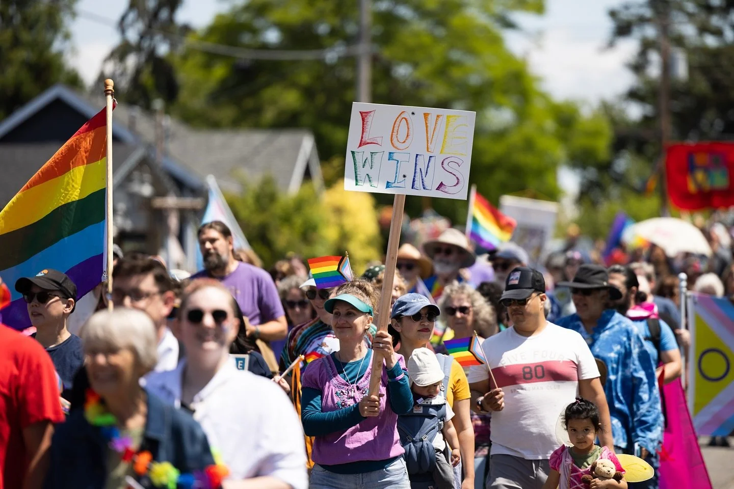 Crowd of people participating in a pride parade, waving rainbow flags and holding signs, including a colorful sign that says 'LOVE WINS', outdoors on a sunny day.