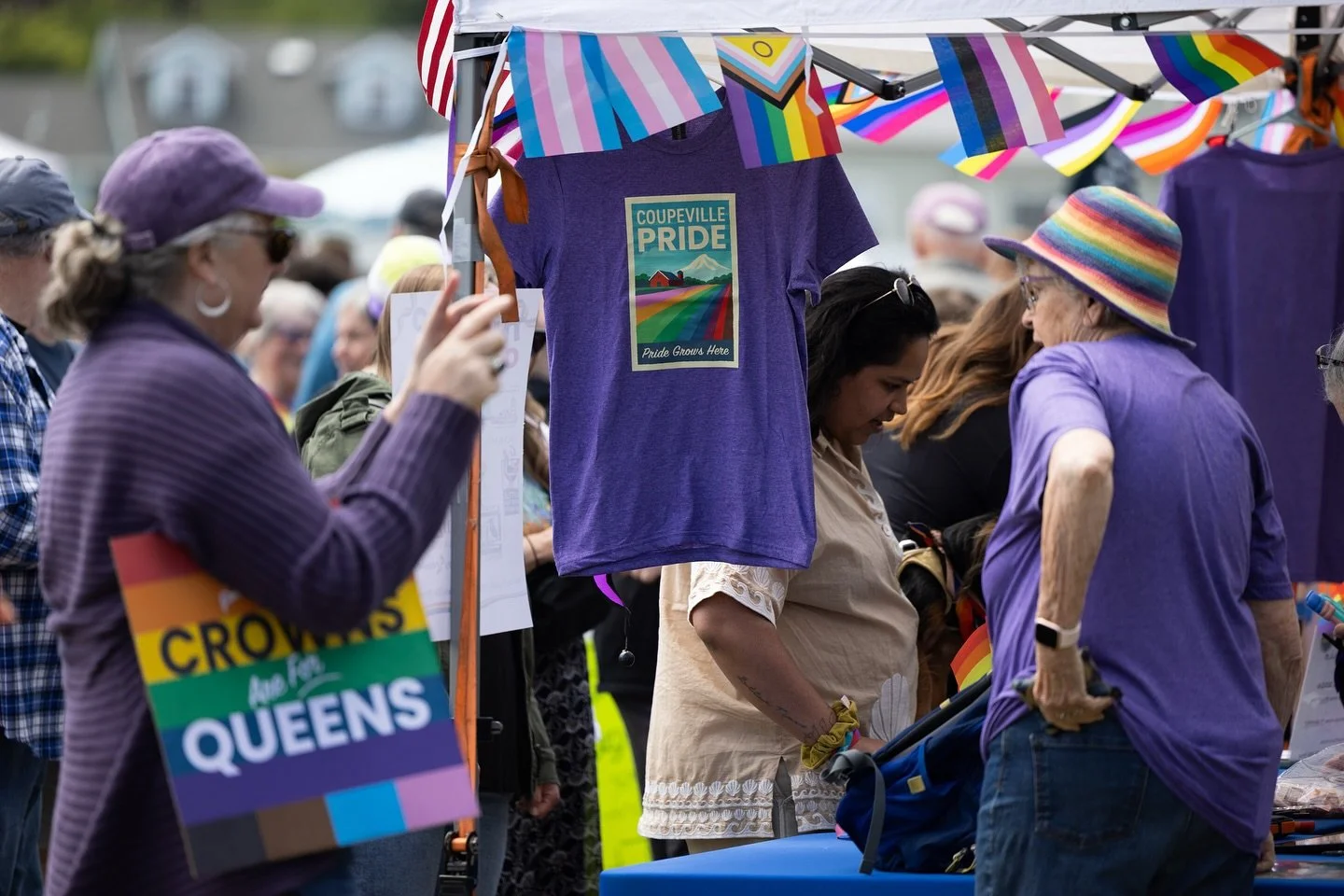 People shopping at an outdoor pride event with rainbow flags and LGBTQ+ themed merchandise, including a purple t-shirt with a pride mountain scene and the words 'Coupeville Pride' and 'Pride Grows Here'.