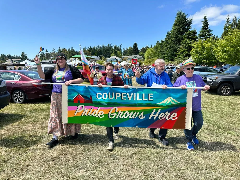 A group of gay people holding a banner that reads "Coupeville Pride Grows Here" at an outdoor pride event, with cars and a crowd in a grassy area under a blue sky with trees in the background.