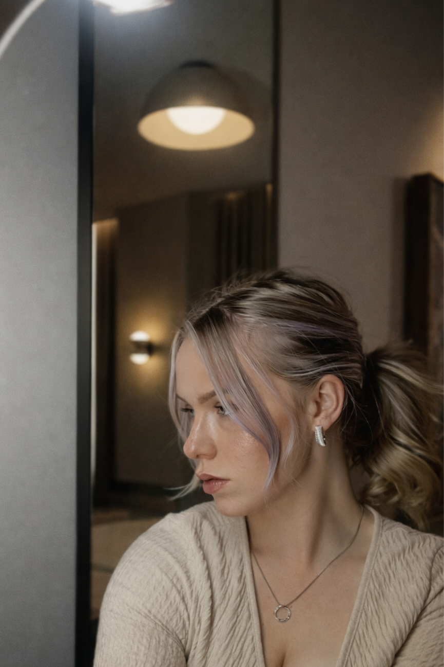 A woman with light-colored hair tied back, wearing earrings and a necklace, looking down with a contemplative expression in a dimly lit room with a mirror and warm lighting.