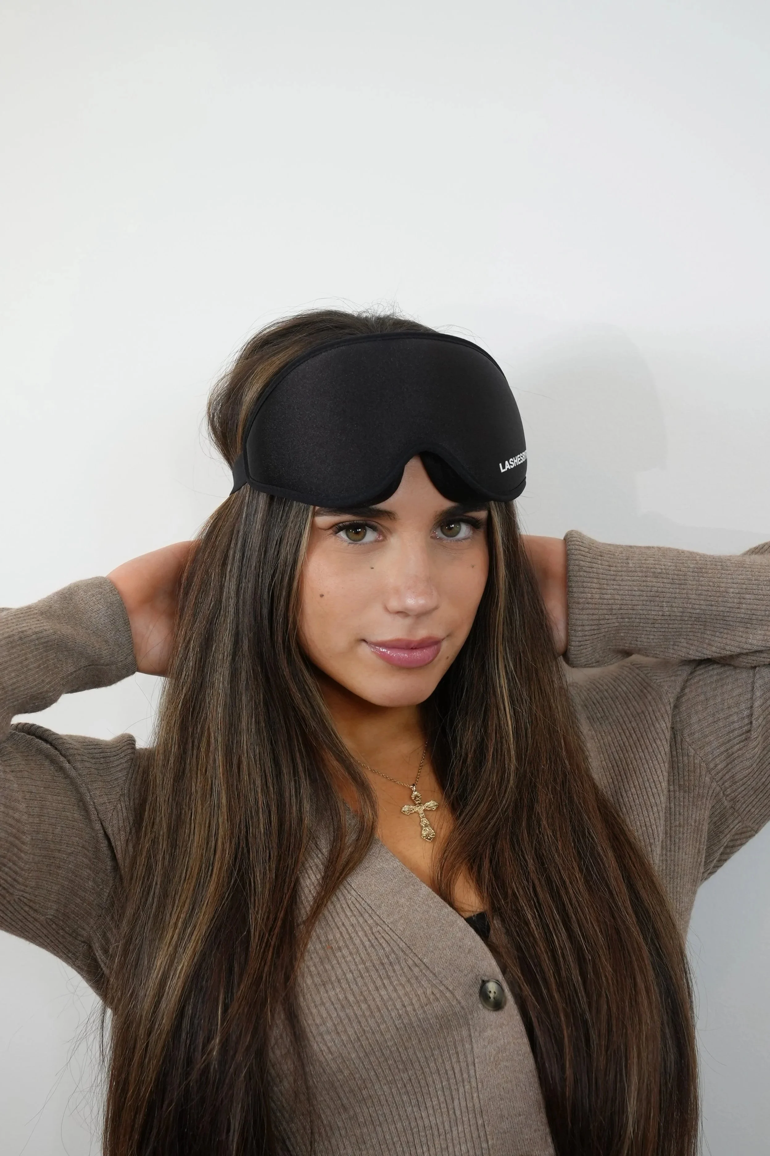 A woman with long brown hair, wearing a tan blazer over a beige top, a gold cross necklace, and a black sleep mask on her forehead, against a plain white background.