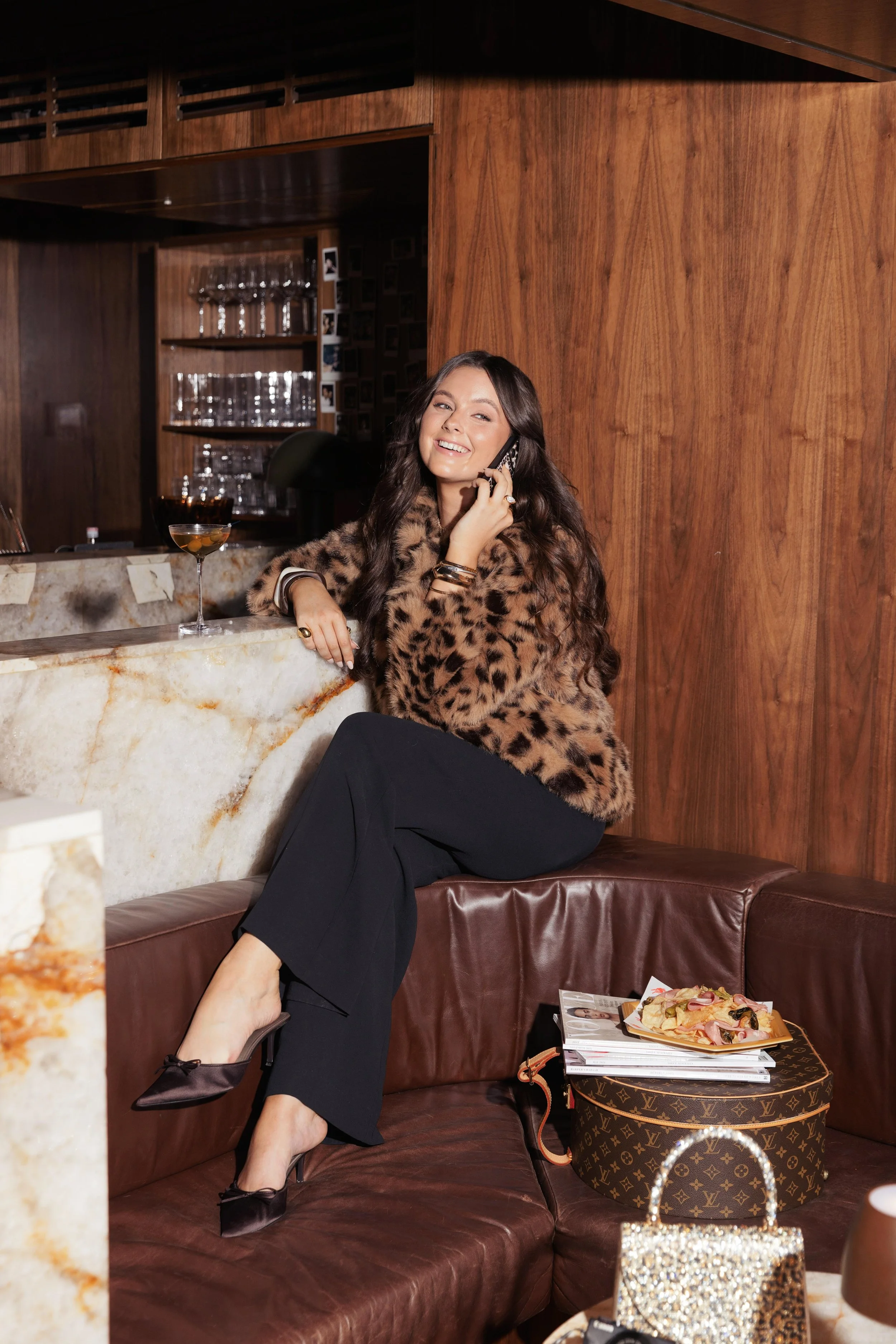 A woman with long dark hair, wearing a leopard-printed coat, black wide-leg trousers, and high heels, sitting on a leather booth in a wood-paneled room. She is smiling while talking on a mobile phone, with a cocktail glass on the marble counter in front of her. There is a stack of magazines, a Louis Vuitton purse, a small glittery handbag, and a plate of food on the table next to her.