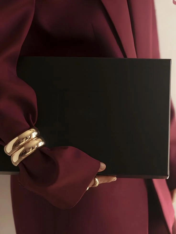 Person dressed in a burgundy suit holding a black box or book, wearing large silver rings and a matching bracelet.