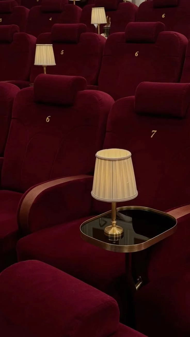 Empty theater seats with numbered labels, plush red velvet upholstery, and small table lamps with beige shades placed on the armrests in a dimly lit room.