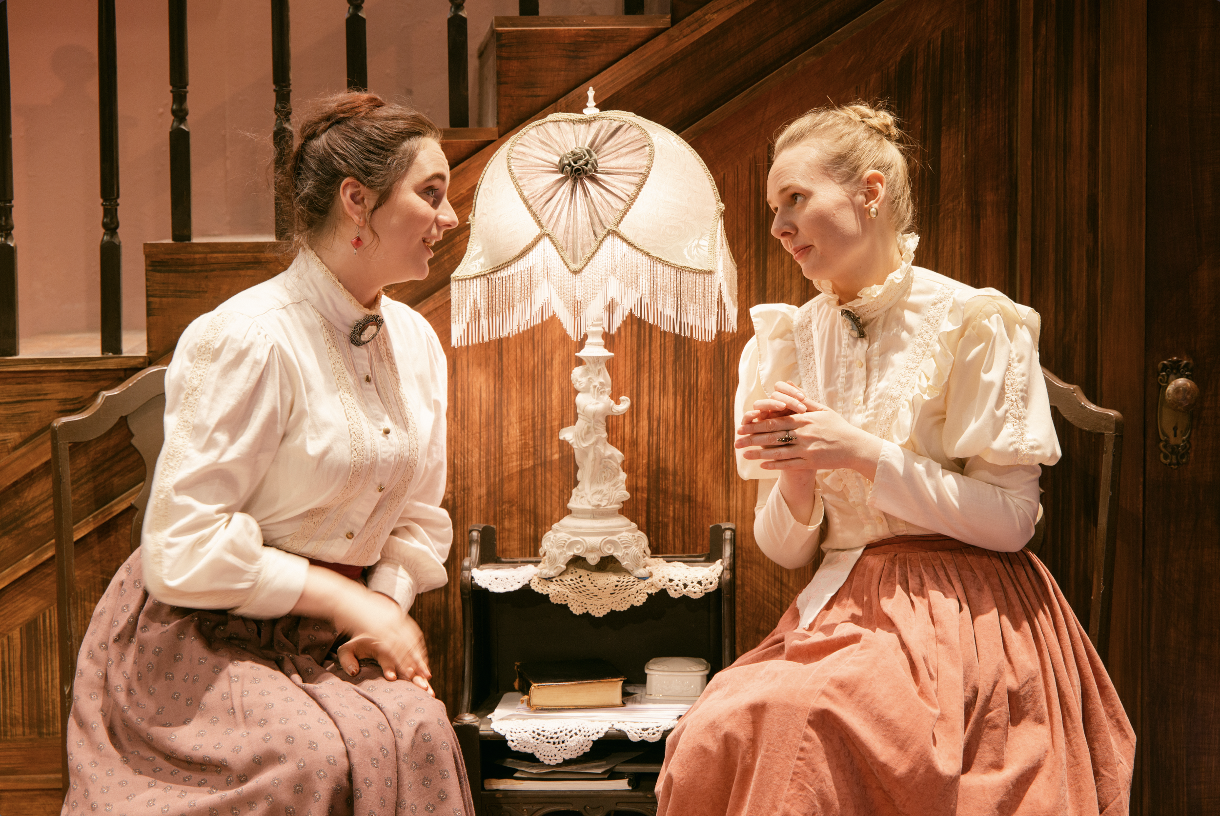 Two women dressed in vintage clothing are sitting indoors next to a ornate lamp with a fringed lampshade. They are engaged in conversation, facing each other, with a wooden staircase behind them.