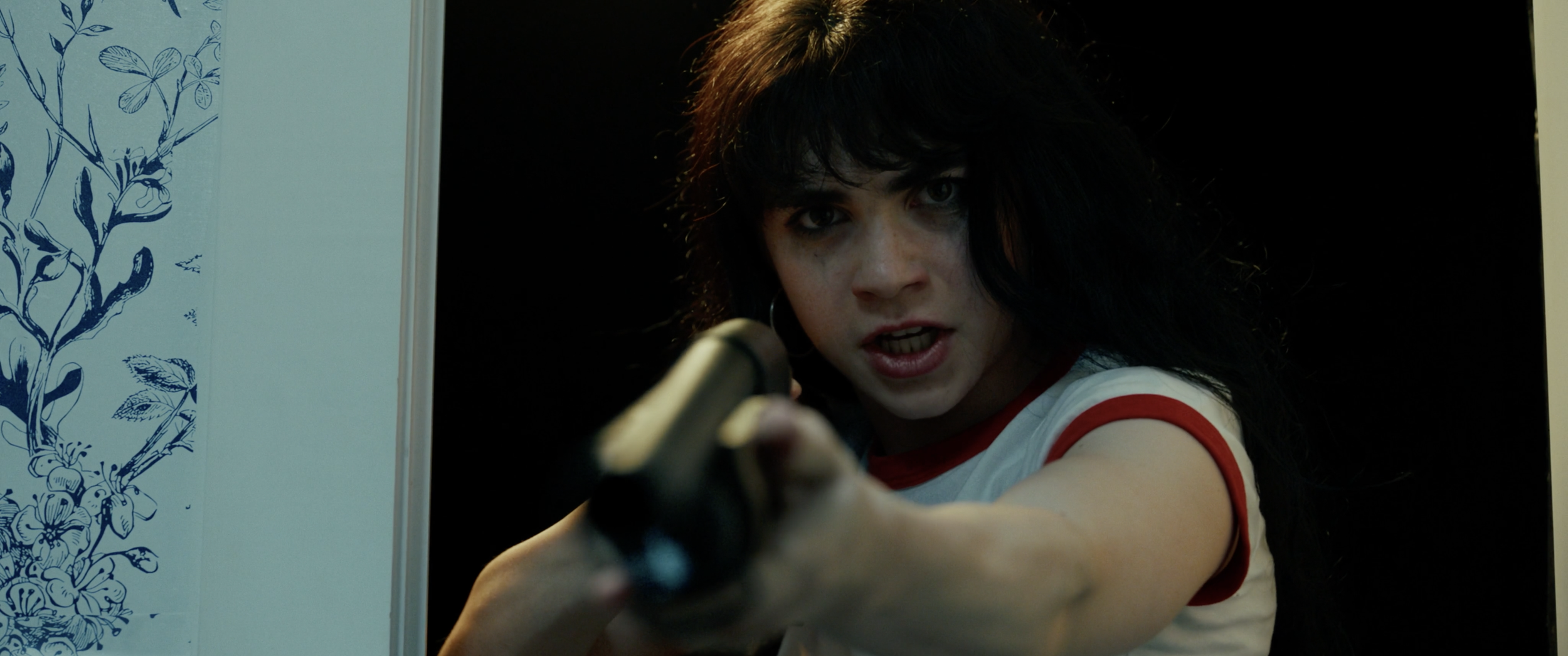 A woman with black hair, wearing a white shirt with red trim, is pointing a gun directly at the camera with a serious expression on her face, in a dark room.