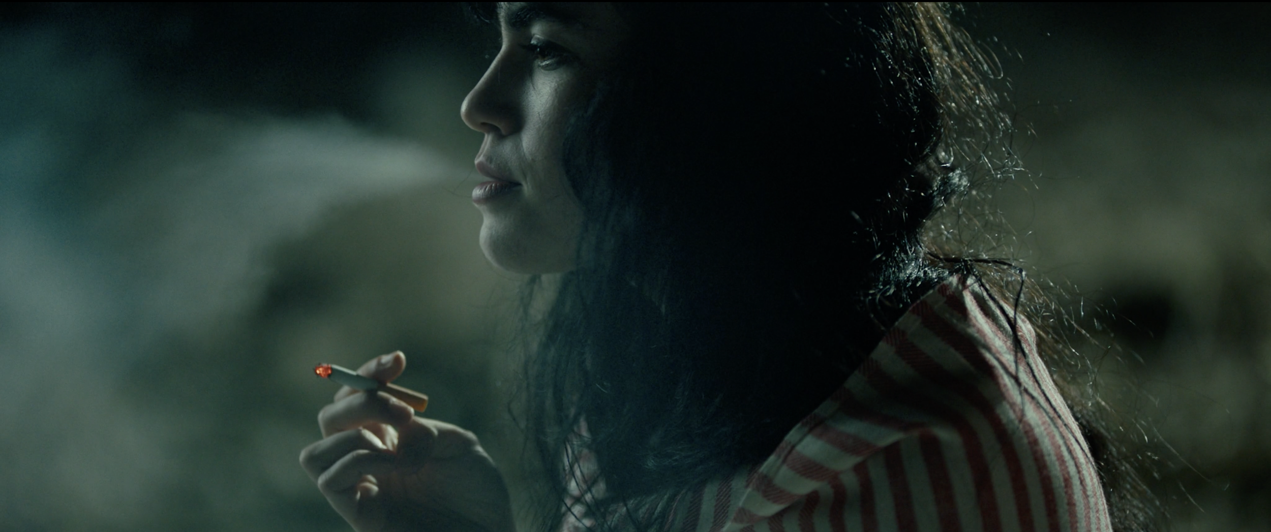 A woman with long dark hair holding a lit cigarette in her right hand, looking to the left side of the image, wearing a red and white striped shirt in a dimly lit outdoor setting.