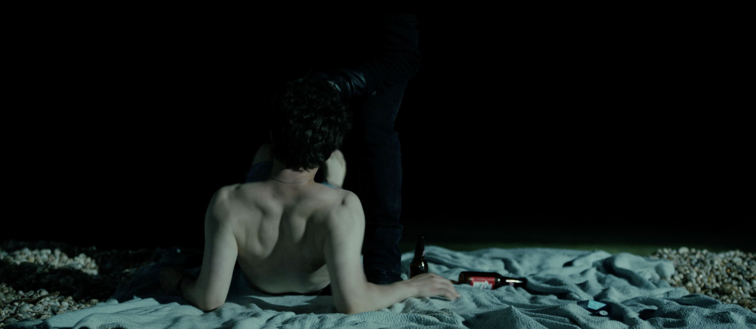 A shirtless man seated on a blanket on a beach at night, with a person standing over him. There are bottles and objects on the blanket.