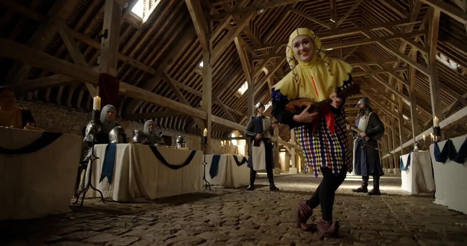 A person in a colorful jester costume performing with a small stringed instrument in a medieval-style banquet hall with long tables, armor-clad participants, candles, and wooden beams.