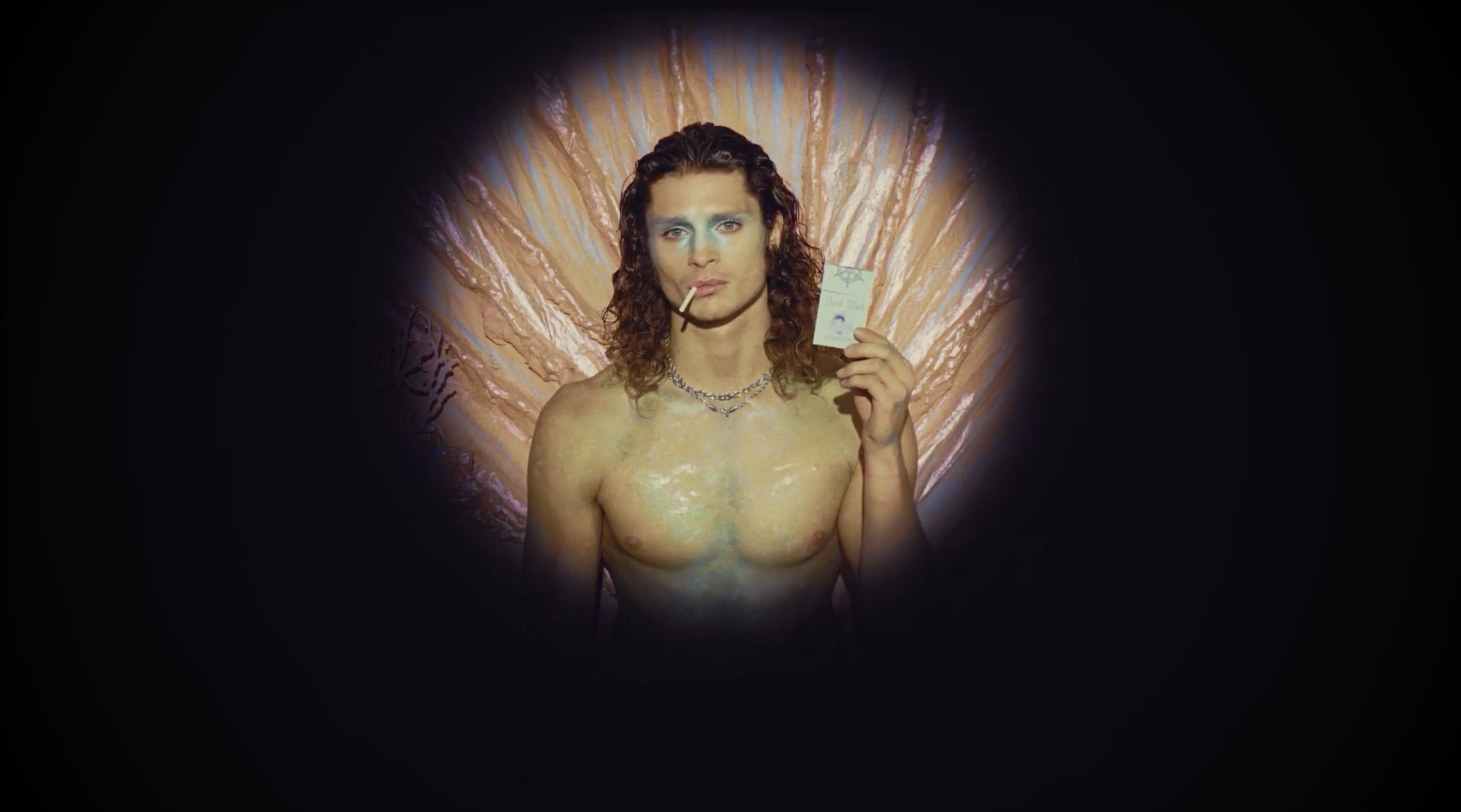 A shirtless man with curly hair and makeup, holding a small note card, standing in front of a colorful, abstract background with a lighthearted and artistic vibe.