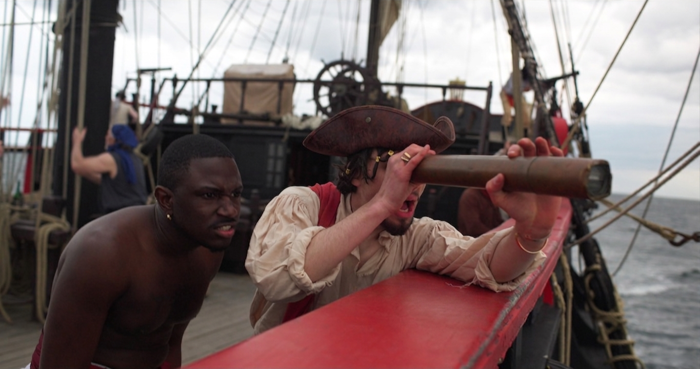 A scene from a ship with two men in historical costumes; one man is looking through a telescope while the other stands behind him, both focused on something in the distance. The ship has rigging and sails, and the background features the ocean and cloudy sky.