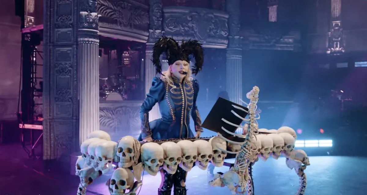 A woman dressed as a DJ with gothic makeup and a feathered headdress, standing behind a DJ booth decorated with skulls and a skeleton, in a dark, ornate venue.