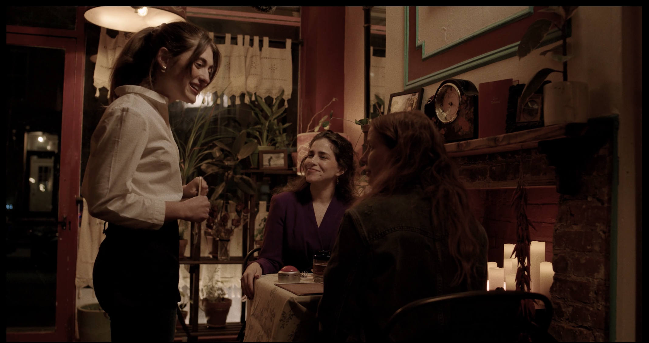 Three women sitting at a table in a cozy, dimly lit restaurant, engaging in conversation. One woman, standing, is holding a small potted plant and smiling. The other two women are seated, one in a purple blazer and the other with long red hair, listening and smiling. The background features warm decor with framed pictures, a clock, plants, and a brick wall.