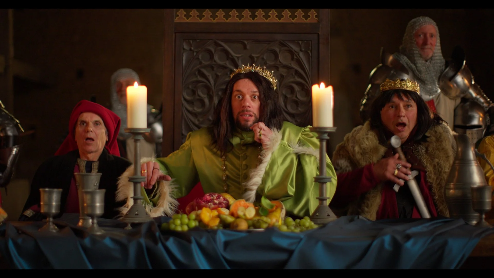 People dressed in costumes sitting at a medieval feast table with candles and fruits, including a man in a green gown with a crown and others with armor and crowns, reacting with surprise.