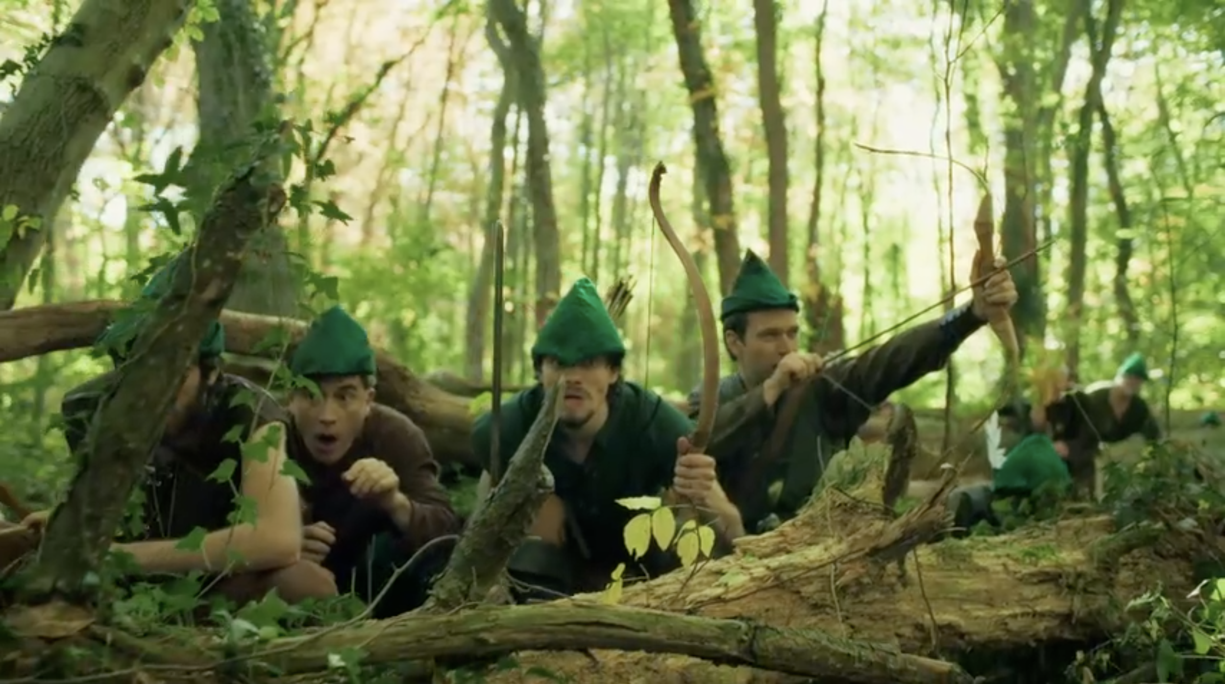 Group of people dressed as elves hiding in a dense forest, with some holding bows and arrows, suggesting a scene from a fantasy or adventure setting.