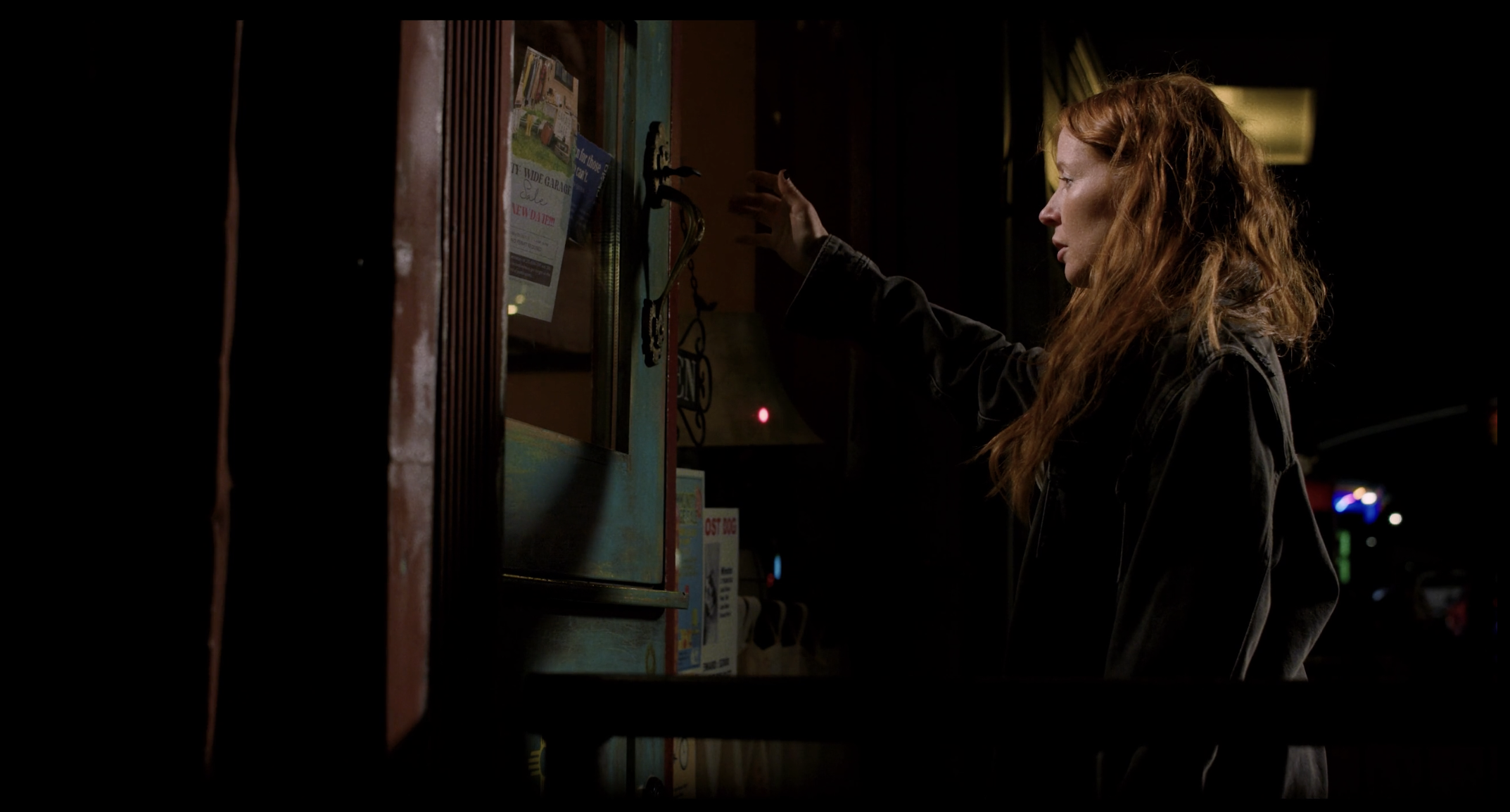 A woman with long red hair wearing a black jacket reaches out to a bulletin board outside at night, with a dark background and few visible lights.