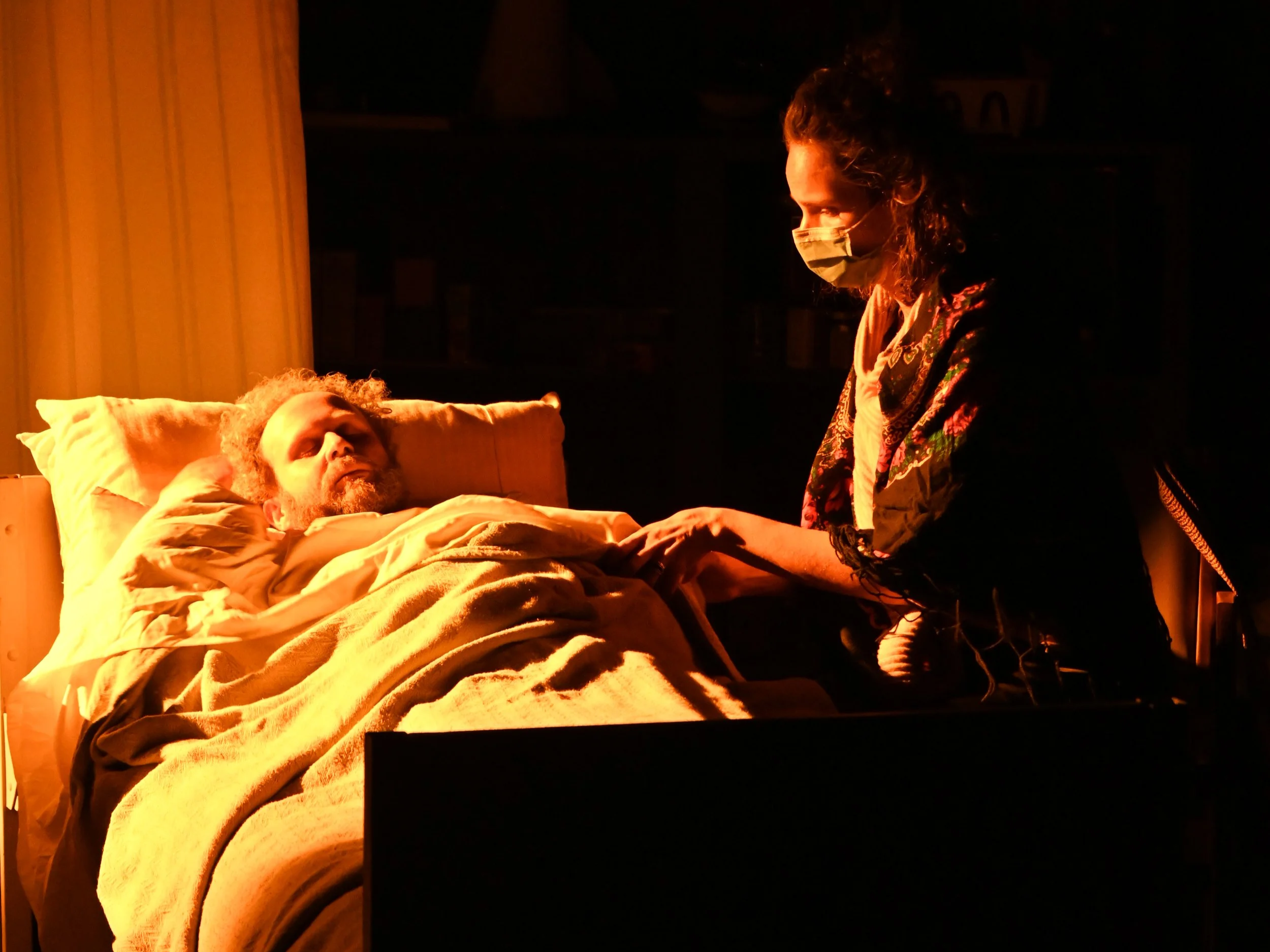 Person lying in bed with curly hair and a beard, wearing a medical mask, holding hands with a person standing beside the bed, in a dimly lit room with a warm glow from a nearby light.