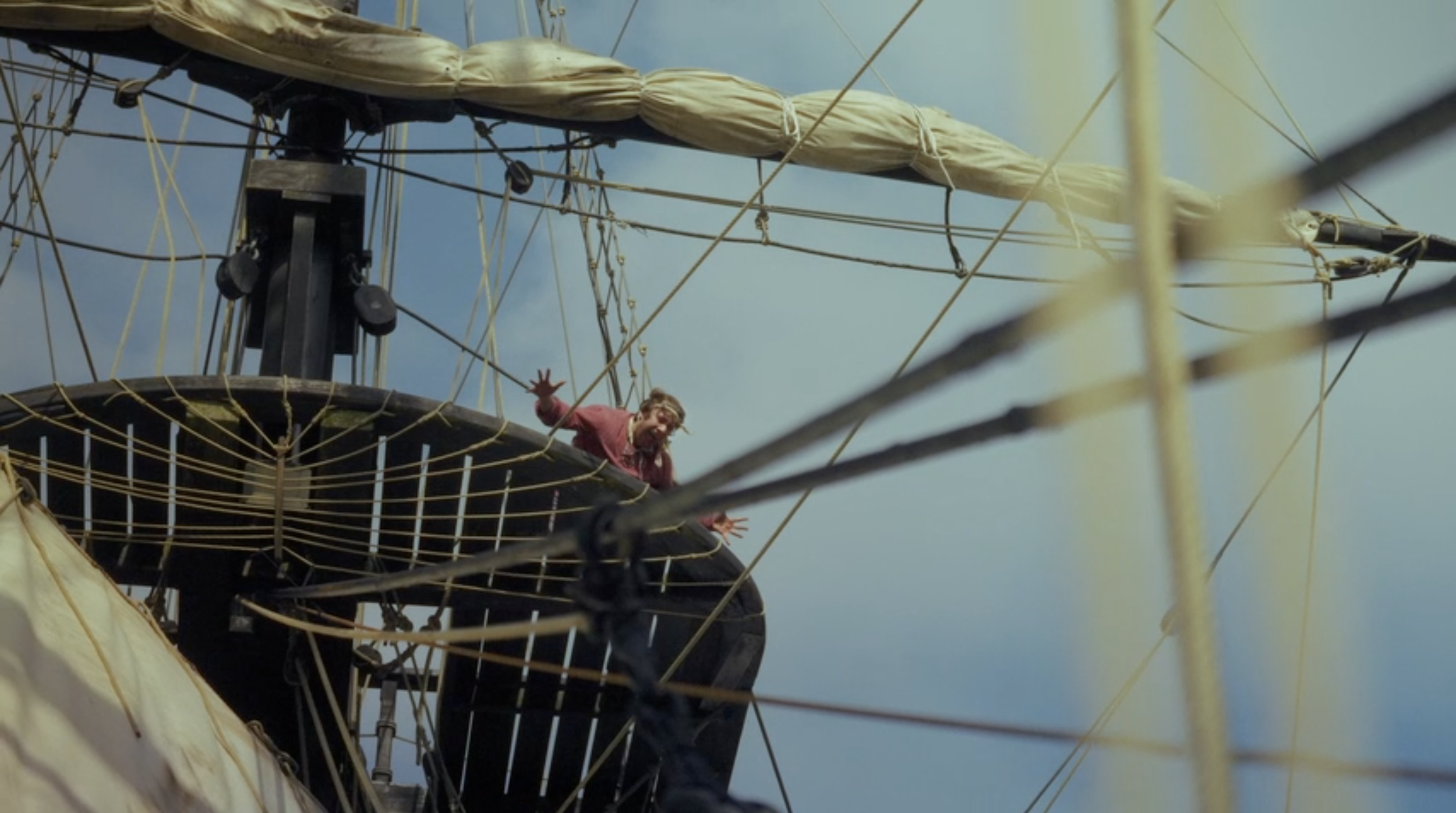 A person with long hair and a beard reaching out with both hands from a ship's crow's nest, surrounded by rigging and sails, against a cloudy sky.