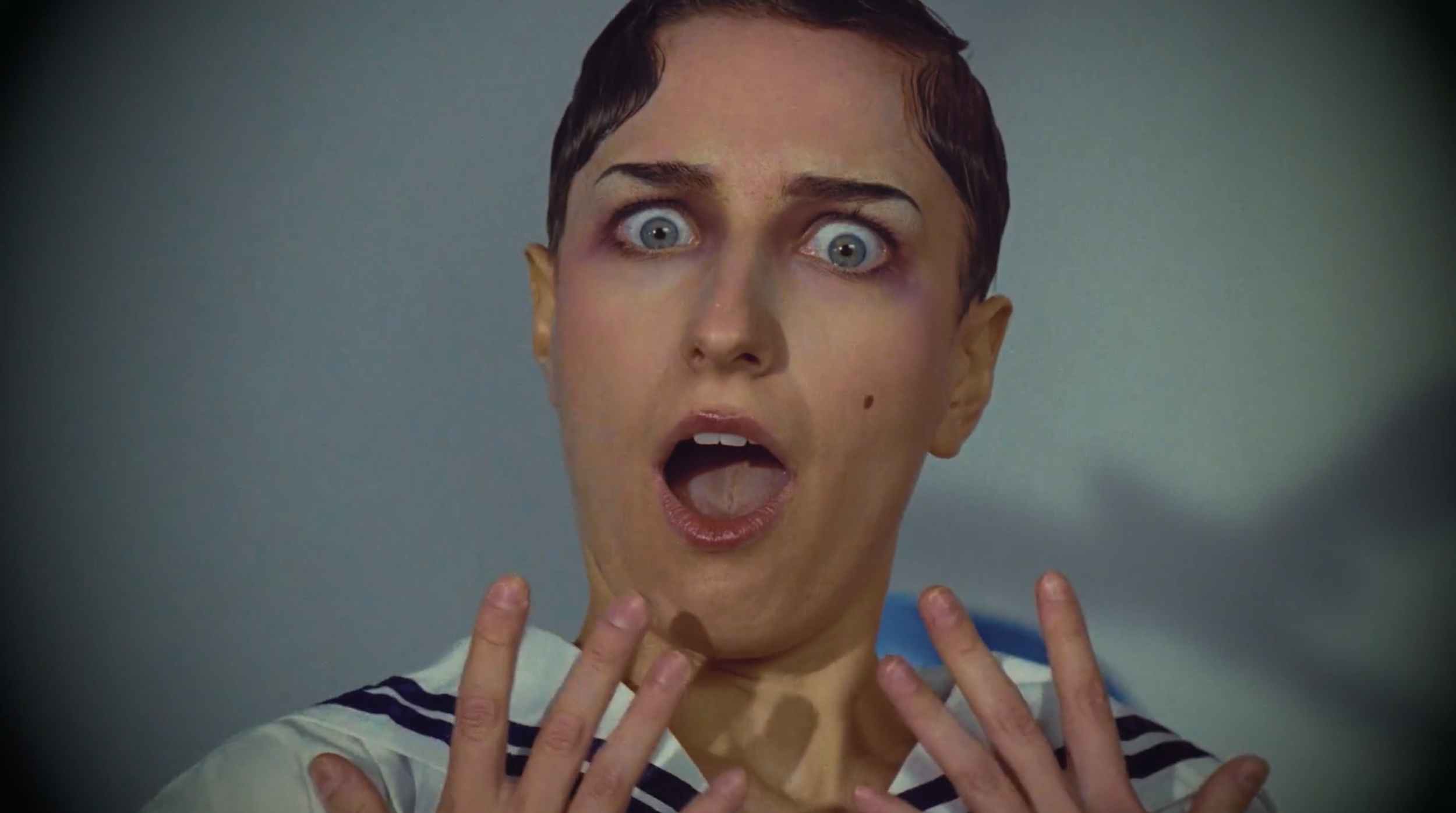 Close-up of a woman with short dark hair and wide eyes, mouth open, with multiple hands touching her face and neck.
