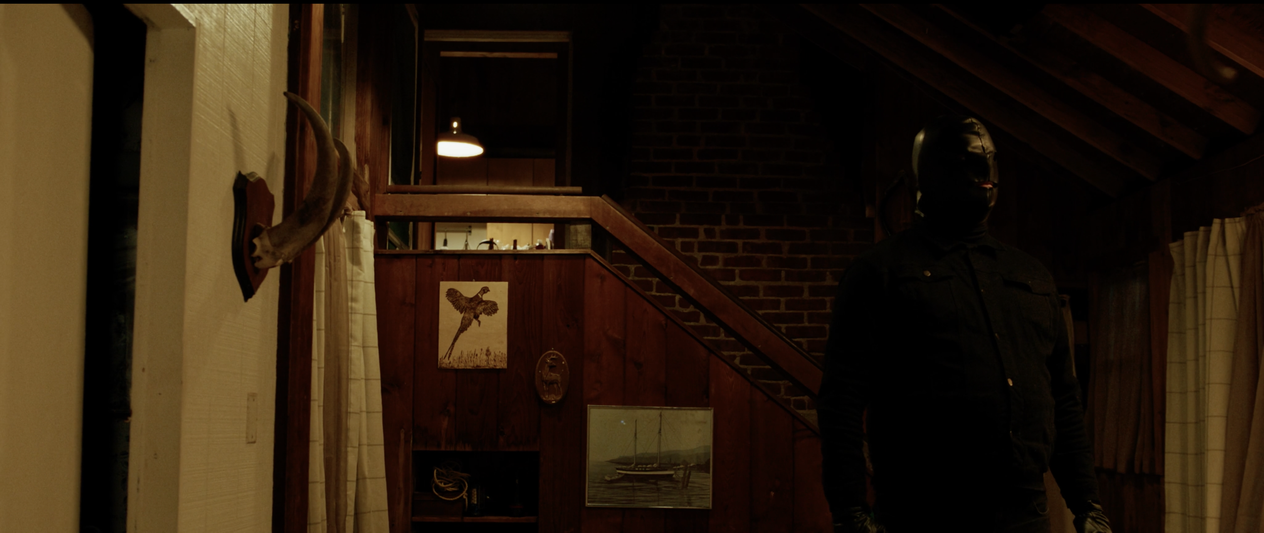 A person wearing a black mask and black jacket standing inside a wood-paneled room. The room has a staircase, a mounted animal head, and pictures on the wall.