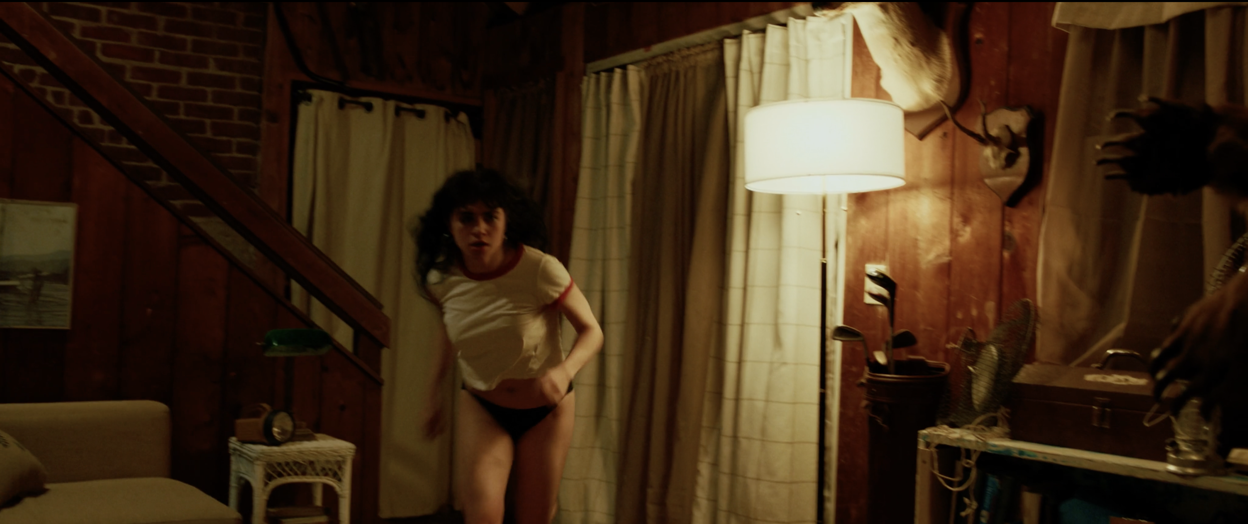 A woman with curly black hair wearing a white t-shirt with red trim and black underwear standing in a dimly lit wood-paneled room, looking towards the camera with a surprised expression.