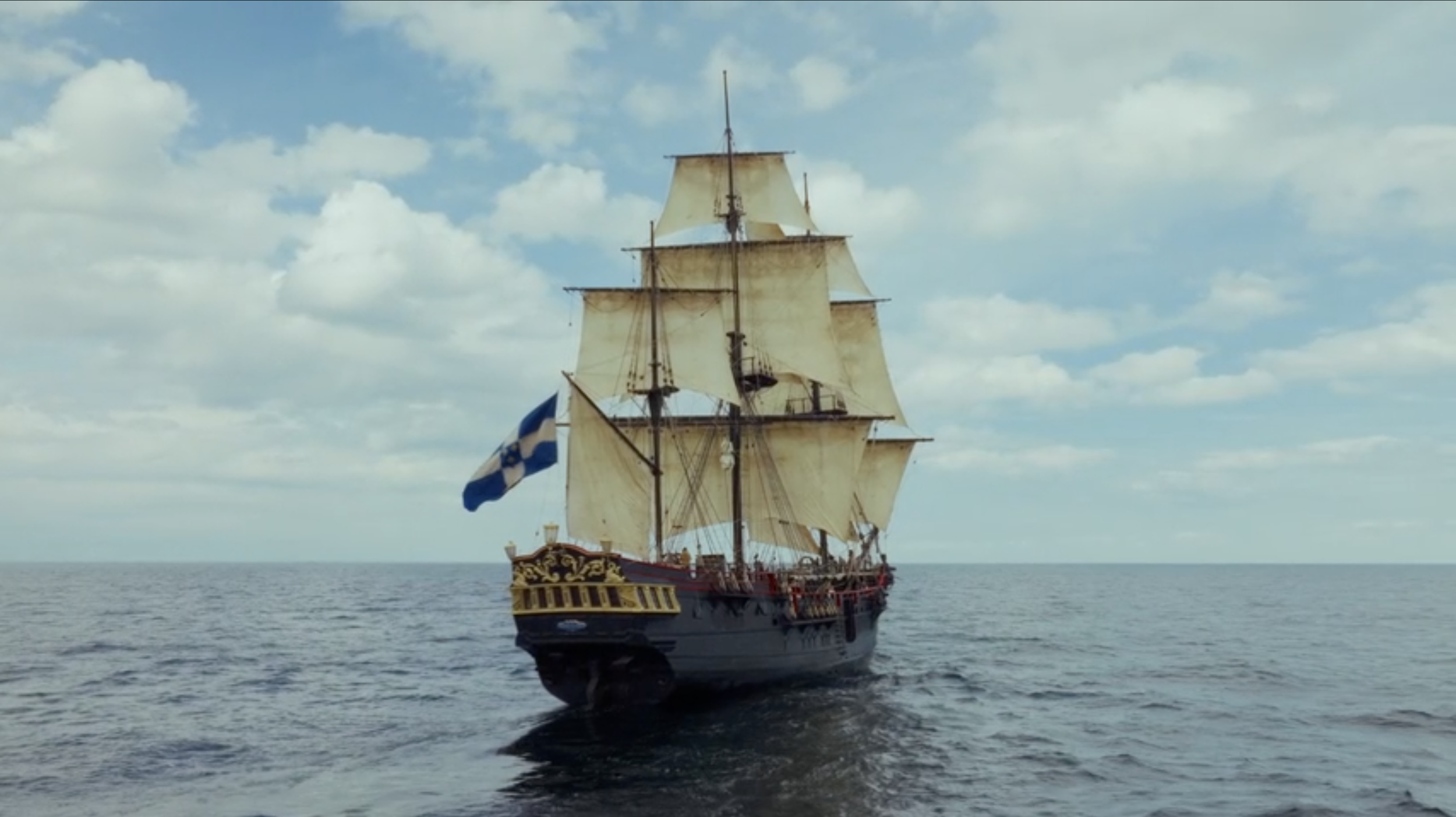 A tall sailing ship with cream-colored sails and a flag featuring a blue cross on a white background, sailing on calm ocean waters under a partly cloudy sky.