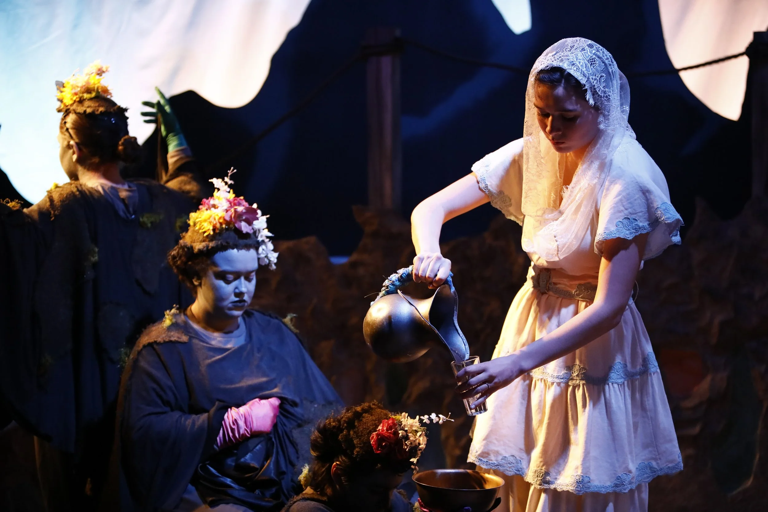 Scene from a theatrical performance featuring women dressed in historical costumes. One woman in a white lace dress pours water from a pitcher, while others, wearing dark costumes with floral headpieces, are engaged in the scene, set against a dark background with a white draped cloth.