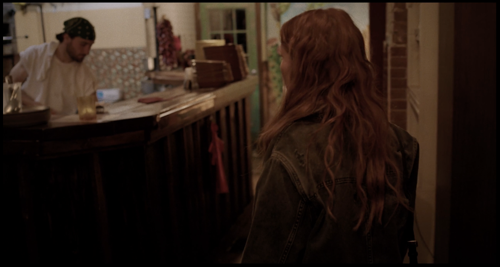 A woman with long, curly red hair stands indoors facing a man behind a wooden bar. The man is wearing a bandana and a white shirt, preparing drinks. The scene has warm, dim lighting with a cozy, rustic interior.