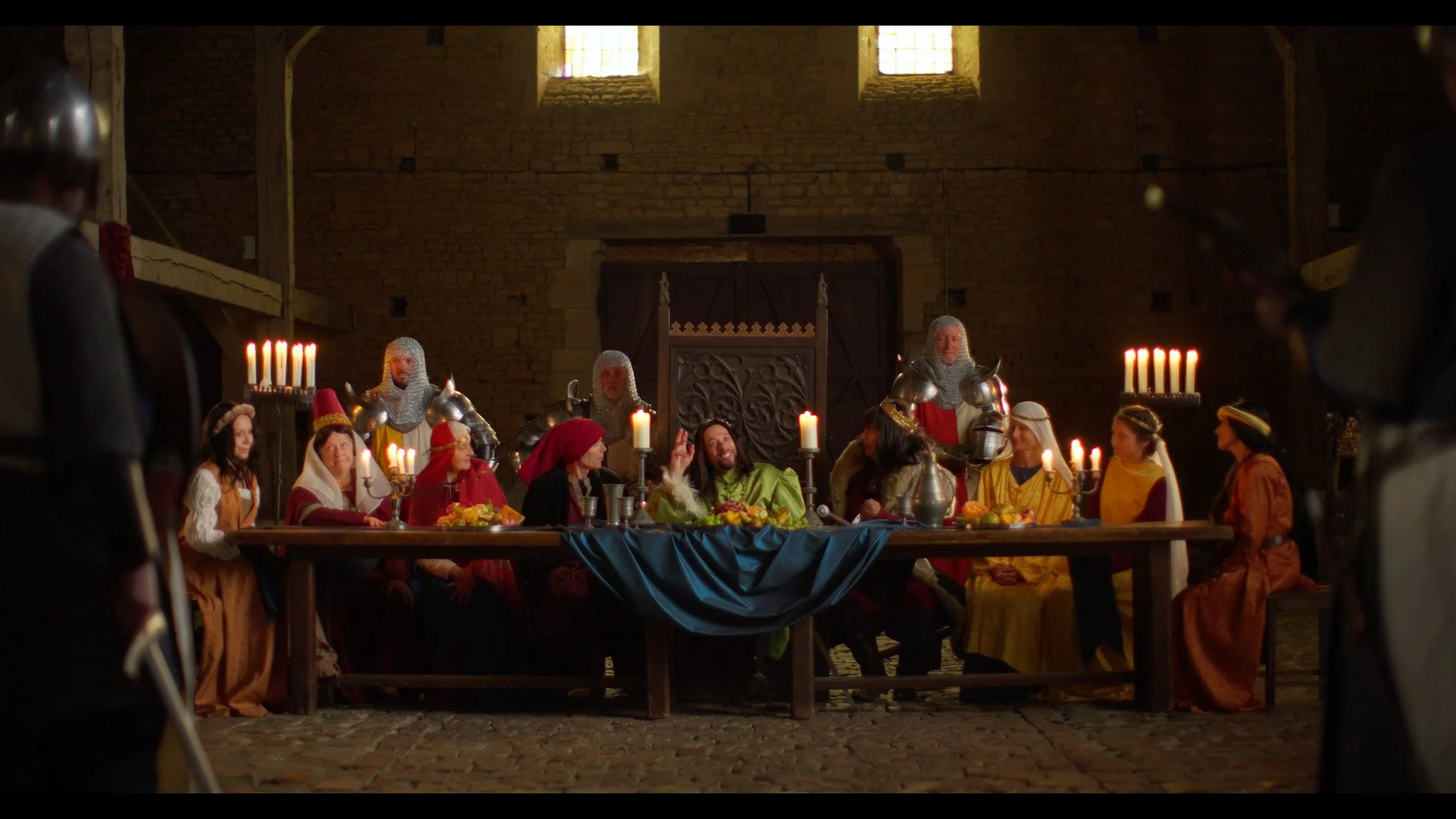 A medieval-style scene of the Last Supper with a group of people dressed in historical costumes seated around a large wooden table, illuminated by candles, inside a stone-walled room with stained glass windows.