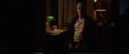 A woman with short blonde hair sitting in a dimly lit room, with a green desk lamp and a kitchen in the background.