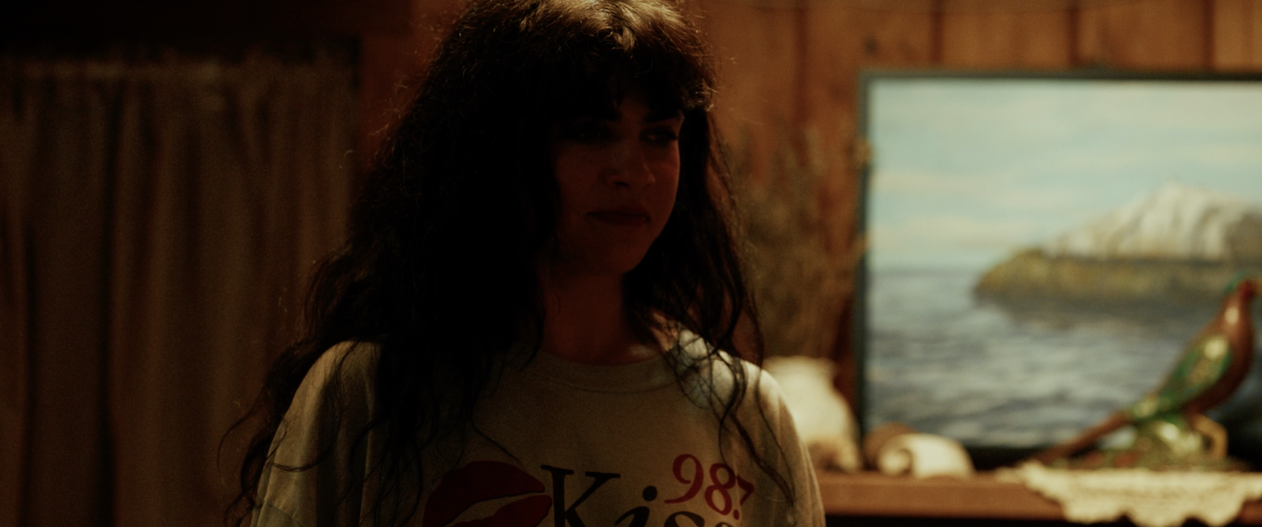 A woman with long, curly dark hair standing indoors in front of a wooden wall. There is a painting of a landscape with water and snow in the background. She is wearing a white T-shirt with red and black text.