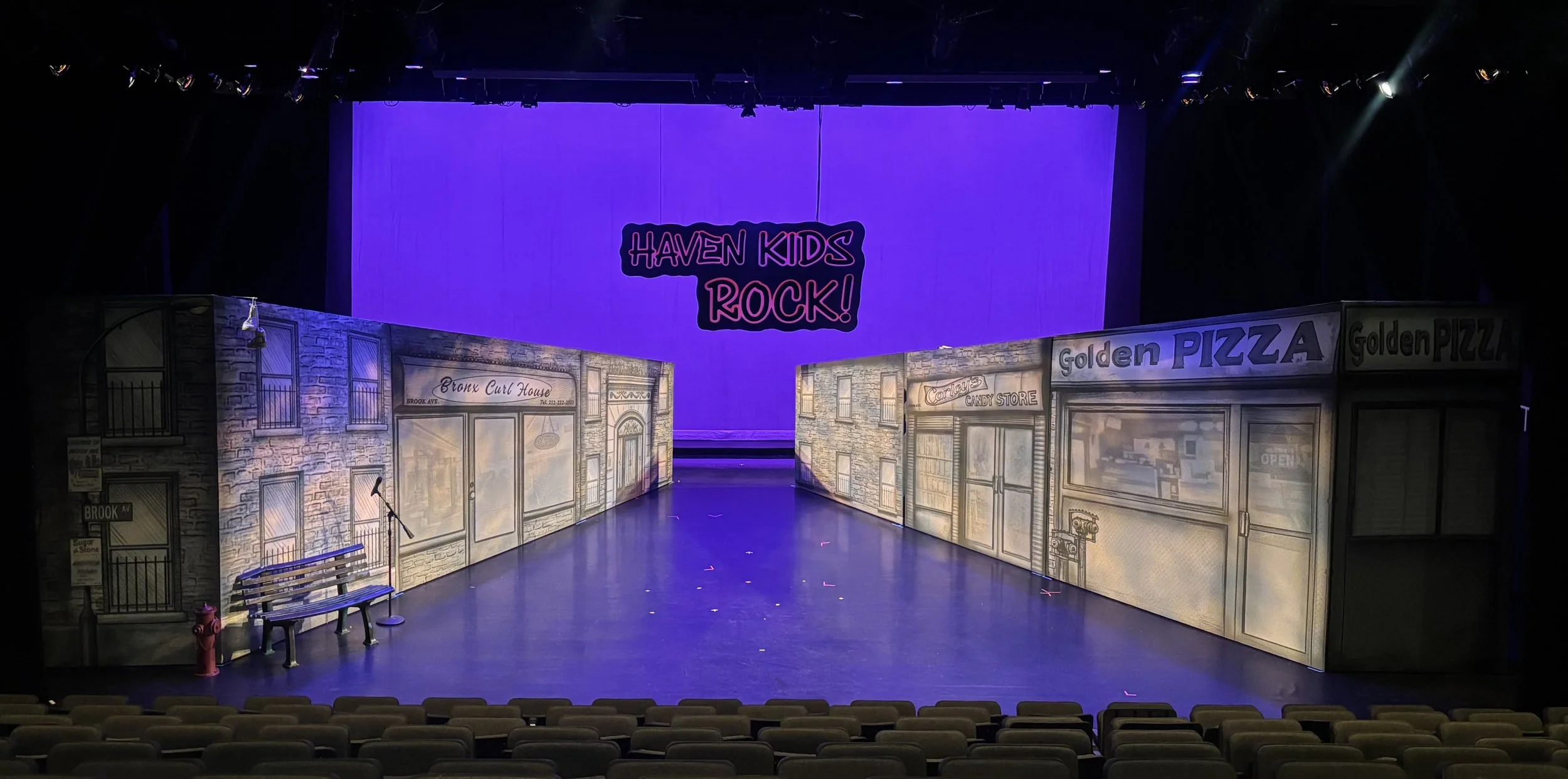 Stage setup with painted storefront facades and a large purple screen overhead displaying the text "HAVEN KIDS ROCK!" in pink. The storefronts include a candy store, a pizza shop, and a Bronx Curl House. There are benches and a microphone on stage, with seating visible in the foreground.