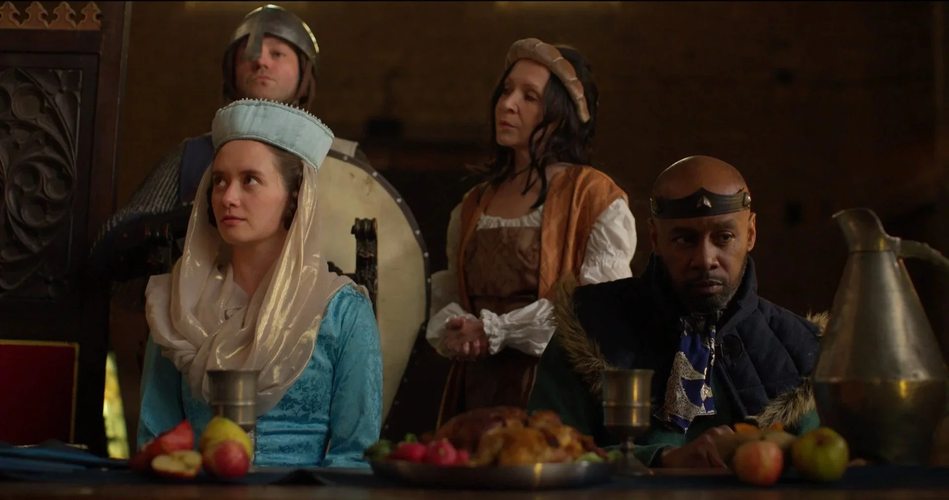 Scene from a medieval feast featuring four people dressed in period costumes, with food and drink on the table, inside a wooden hall.