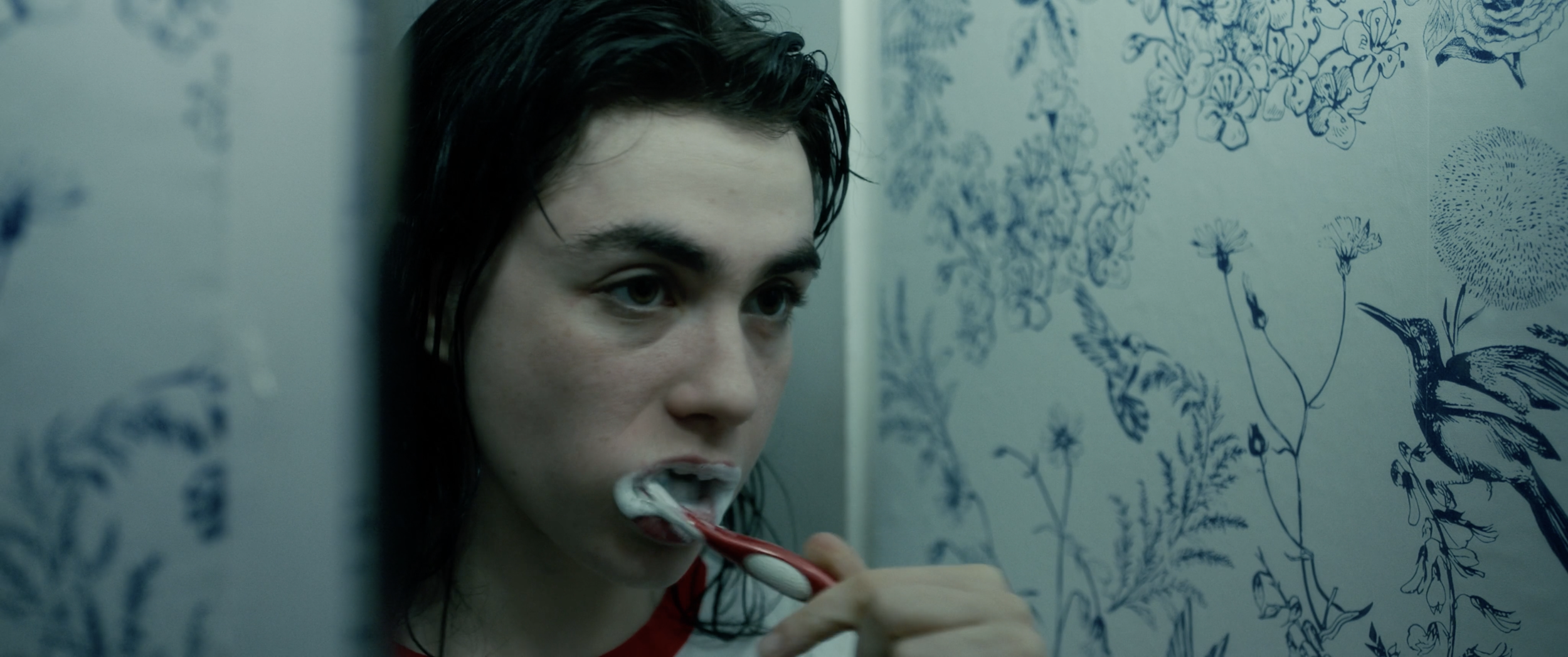 A young woman with wet hair brushing her teeth in a bathroom with blue floral wallpaper.