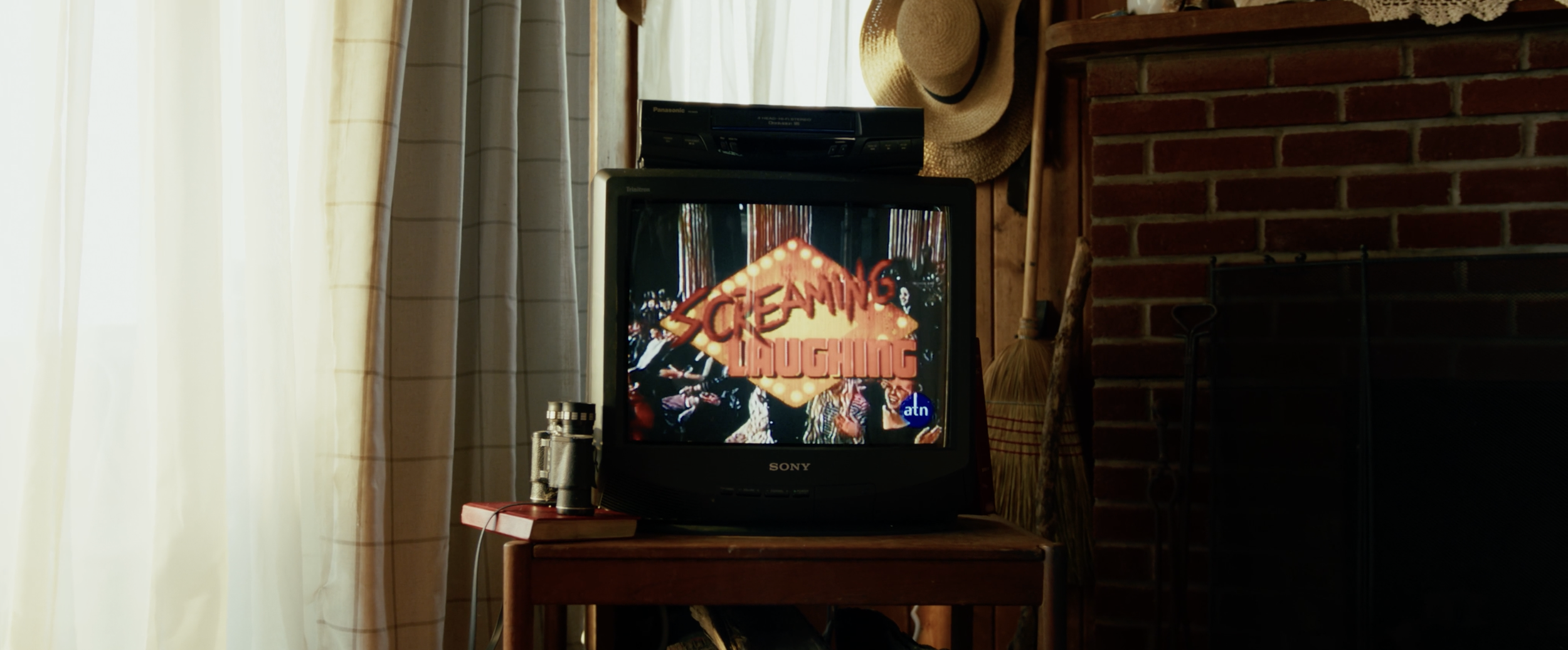 A vintage television displaying the title screen of the movie 'Screaming Through' in an room with a brick fireplace, beige curtains, and a straw hat hanging on the wall.
