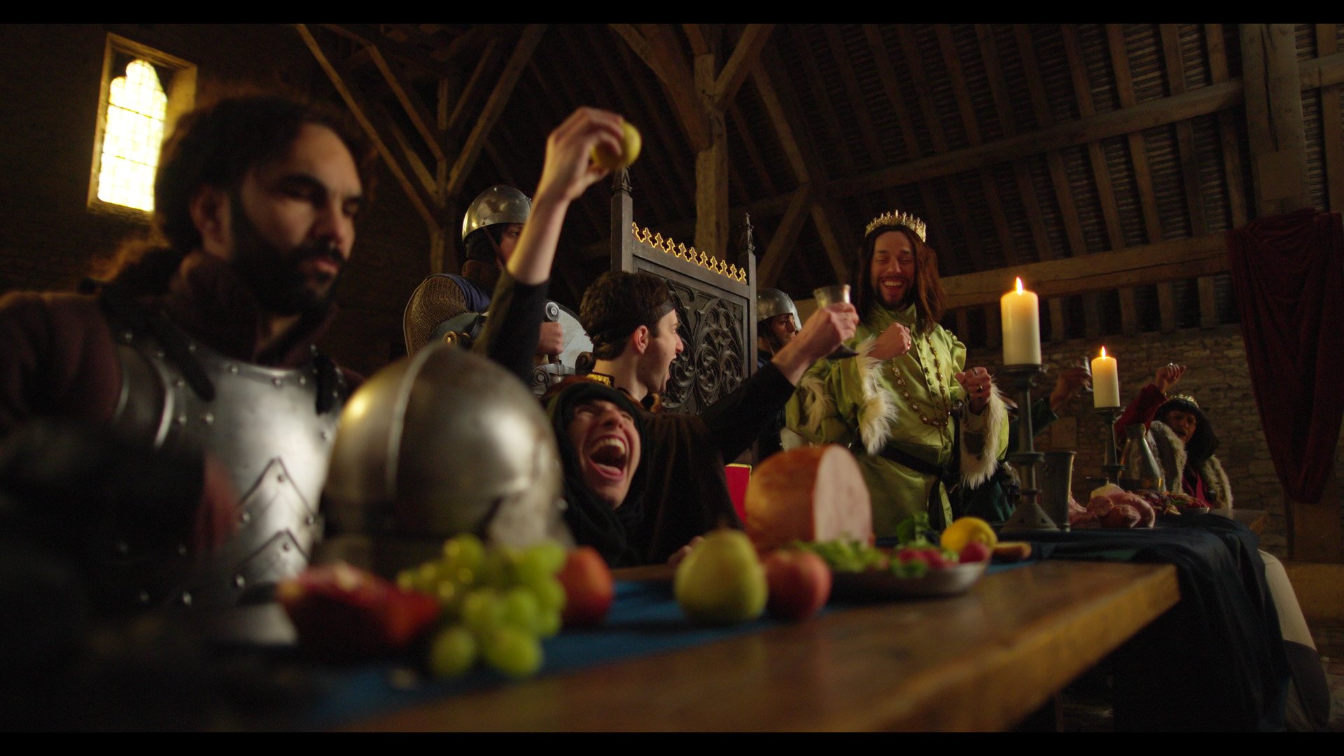 People dressed in medieval costumes celebrating at a banquet inside a castle hall with candles, food, and decorations.