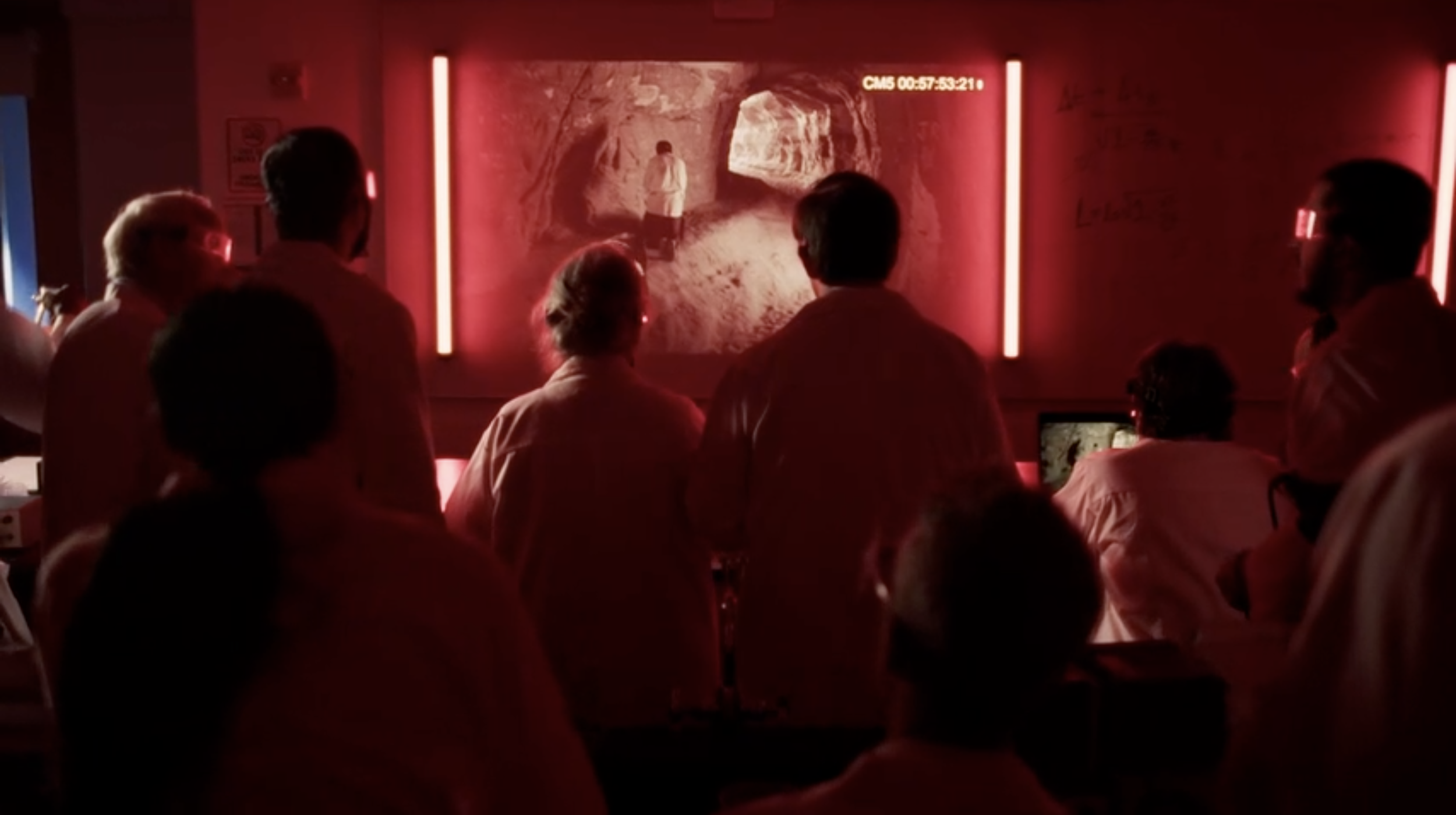 People gathered in a dark room watching a large projected image of a person in a tunnel or cave with red neon lights surrounding the display.