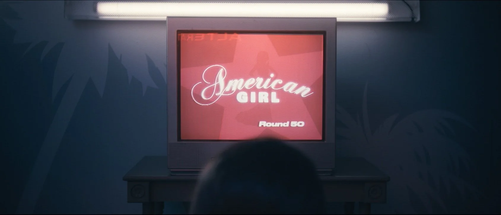 A television screen displaying the title 'American Girl' with 'Round 50' written below, placed on a dark table against a blue wall with faint palm tree shadows.
