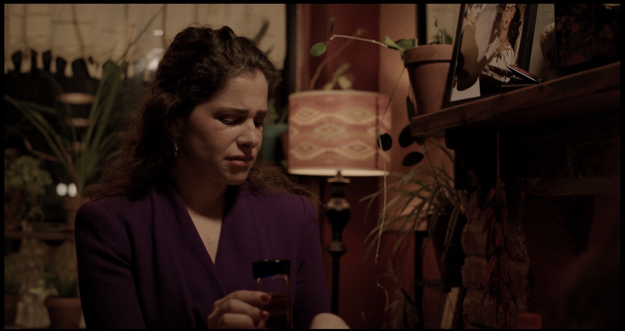 A woman with wavy brown hair wearing a purple blazer looking at her phone in a cozy, warmly lit room decorated with potted plants, a framed picture, and a patterned lamp.