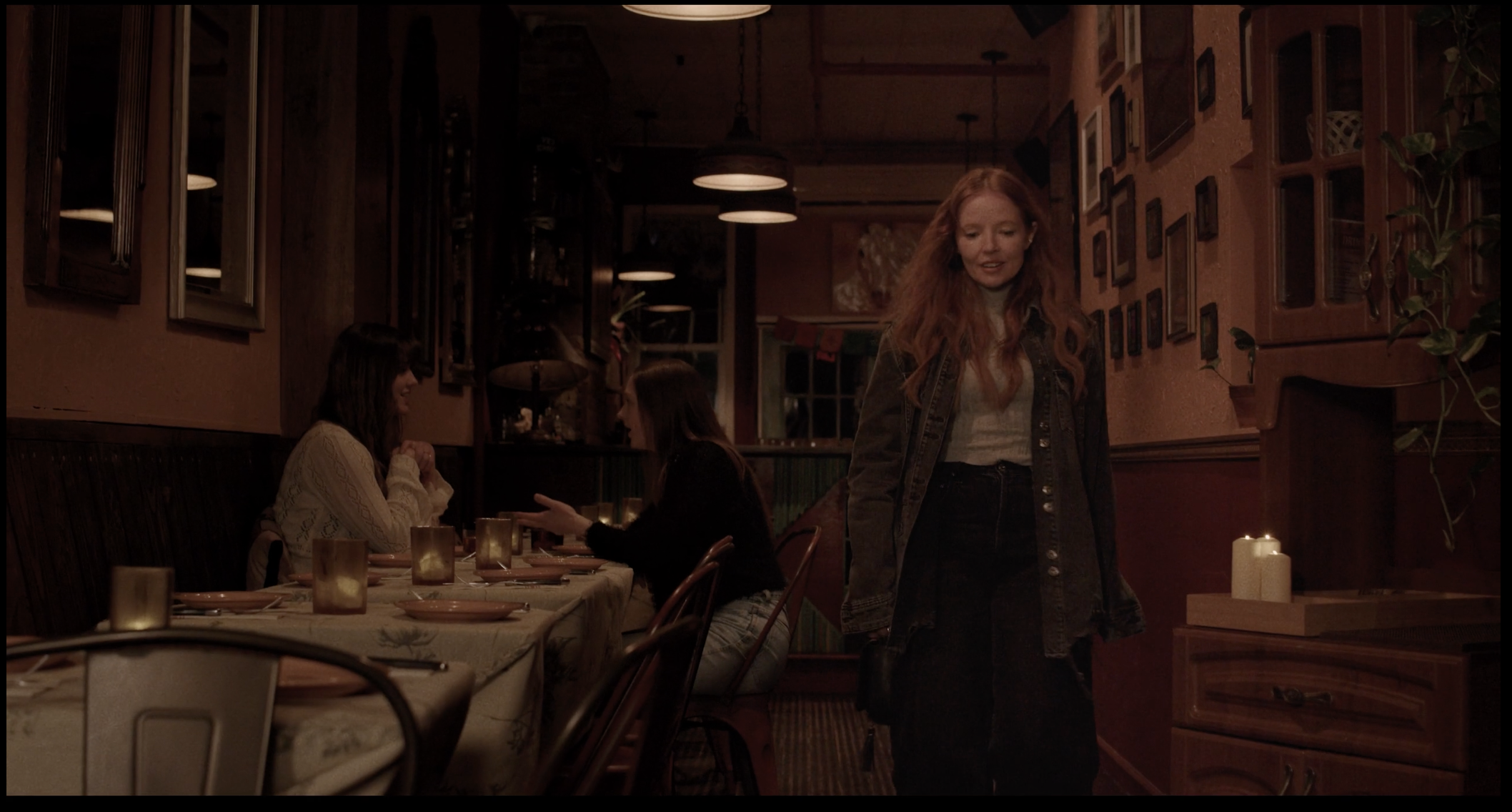 A woman with red hair and a denim jacket standing inside a dimly lit restaurant with people seated at tables, chatting, and candlelit decor.