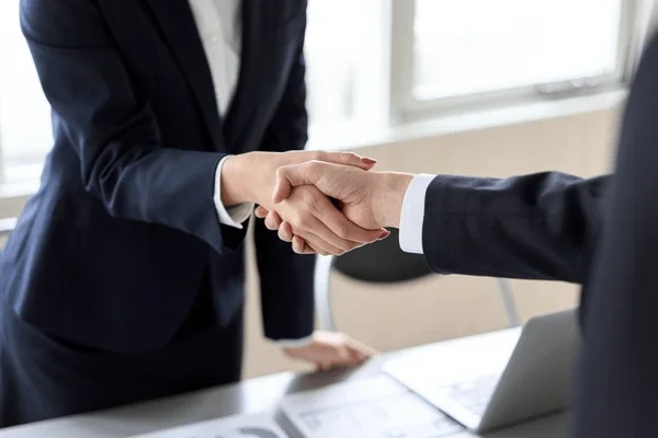 Two people shaking hands in a professional setting with a table and laptop in the background.