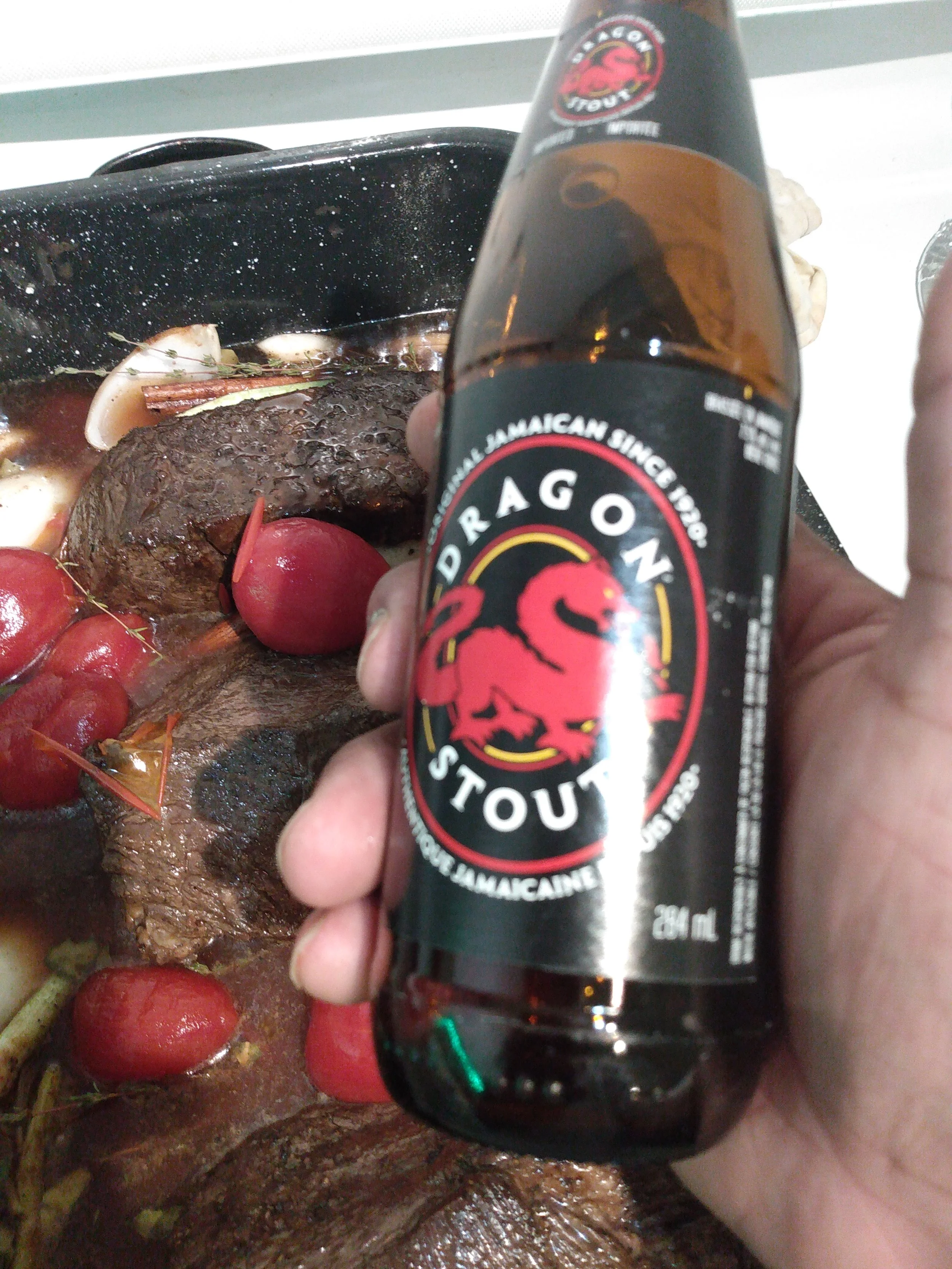 A hand holding a bottle of Dragon Stout beer in front of a pot of cooked meat and vegetables.