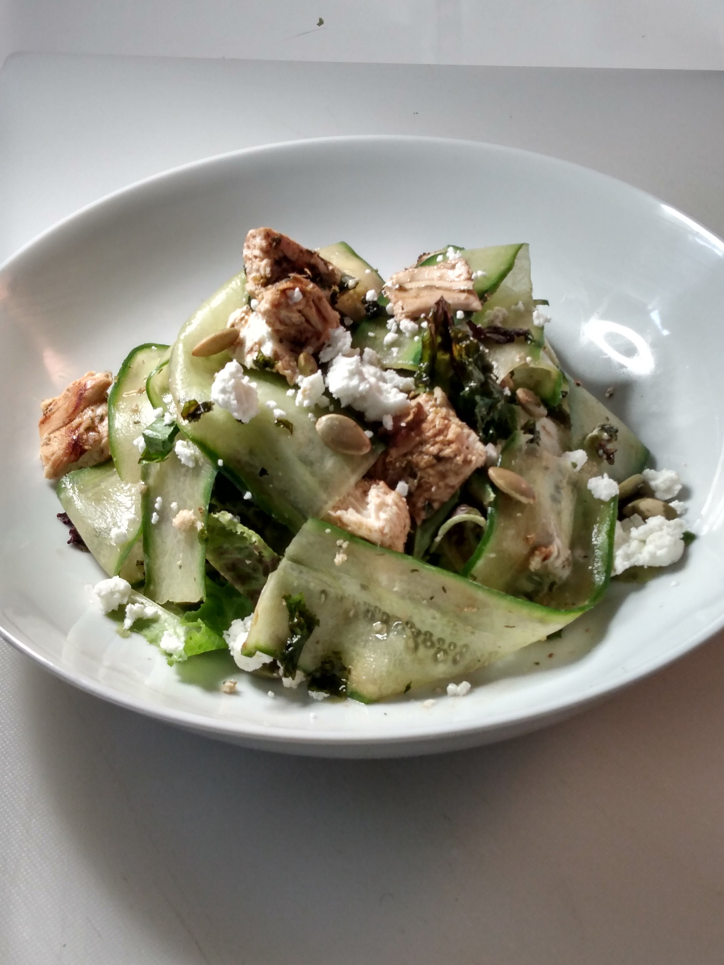 Salad with cucumber ribbons, crumbled cheese, chunks of chicken, sunflower seeds, and mixed greens in a white bowl.