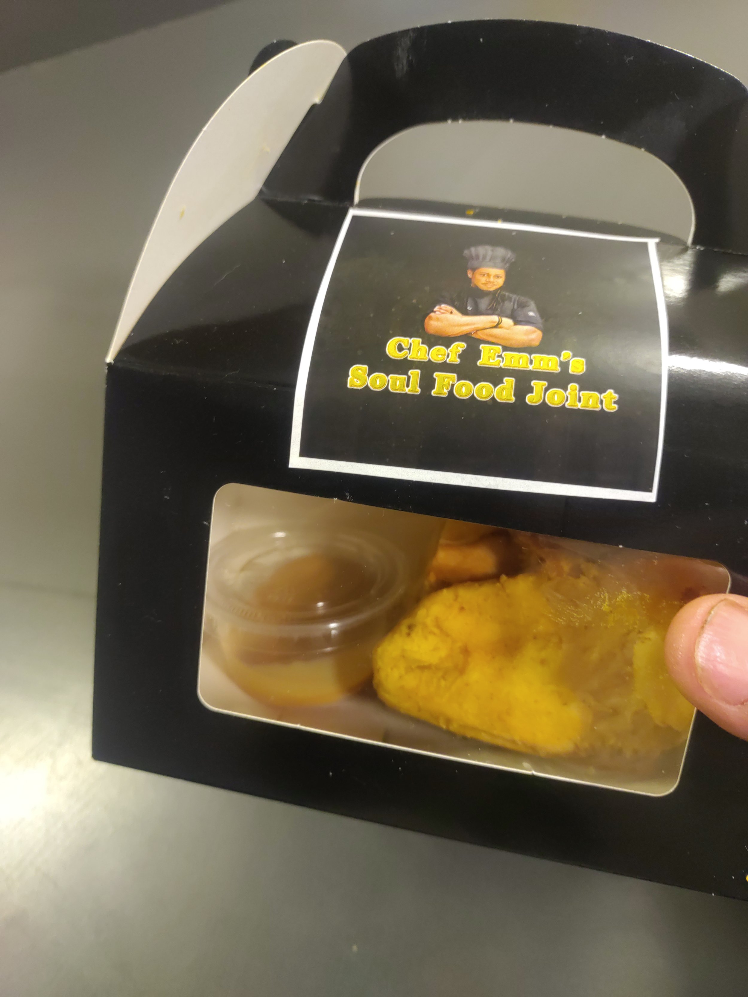 Black food container with a handle and a clear plastic window displaying a piece of fried chicken and a small container of dipping sauce, with a label featuring a chef in a black uniform and hat, and the text 'Chef Emm's Soul Food Joint'.