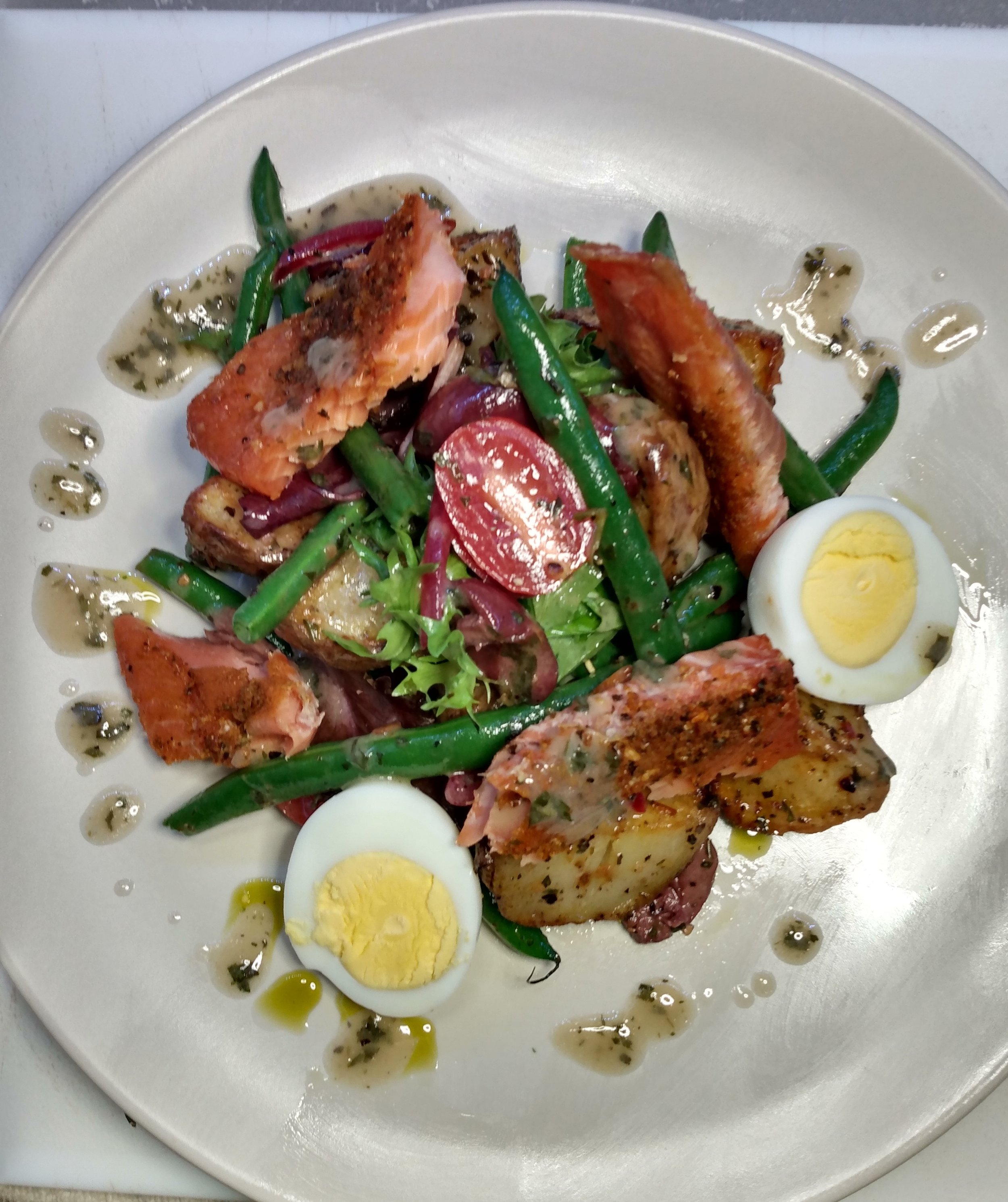 Salad with boiled eggs, green beans, cherry tomatoes, roasted potatoes, and smoked trout, garnished with herbs and dressing.