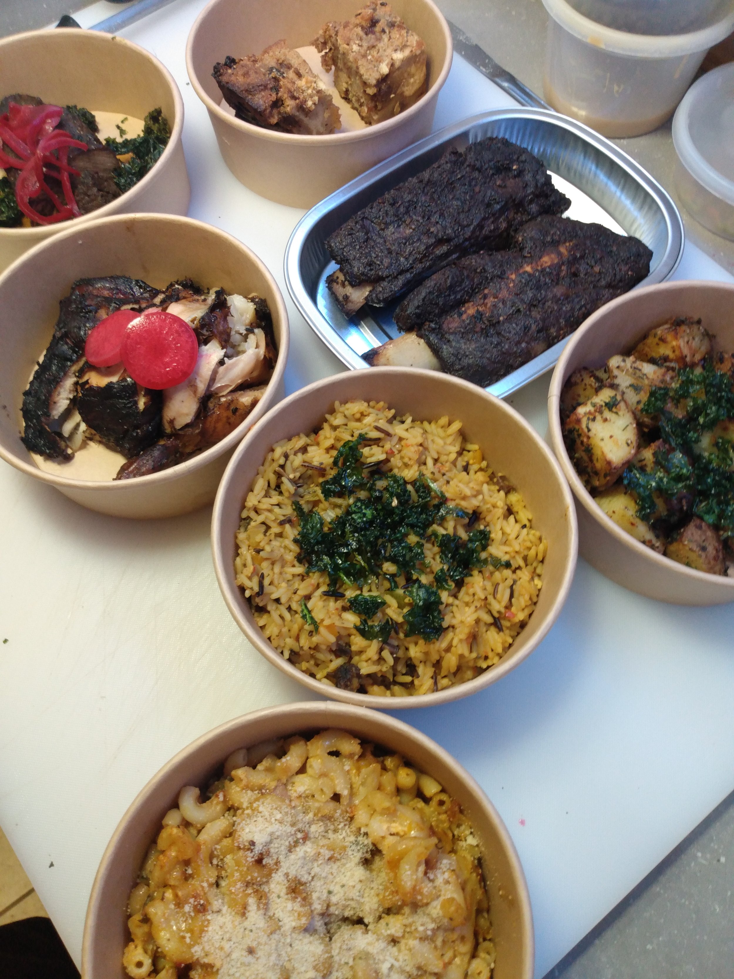 Assorted dishes of grilled ribs, rice, pasta, roasted chicken, and vegetables in bowls with a metal tray of smoked ribs in the center.