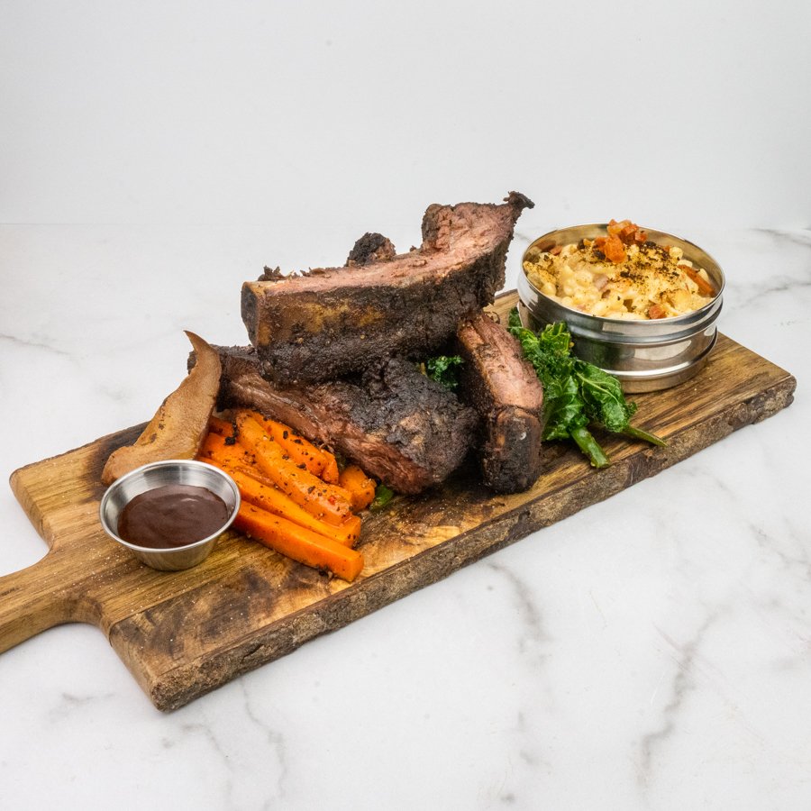 Beef back ribs with sauteed carrots, green leafy vegetables, smoked pear, mac & cheese, and sauce on a wooden serving board.