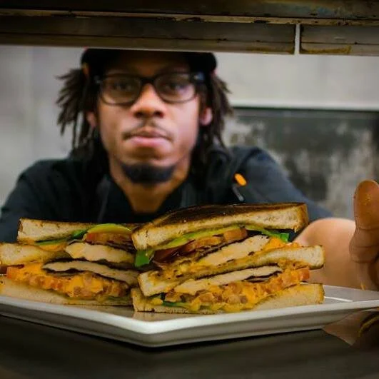 A person with glasses and dreadlocks looking at a plate of three large sandwich stacks with toasted bread, chicken, lettuce, and sauce.